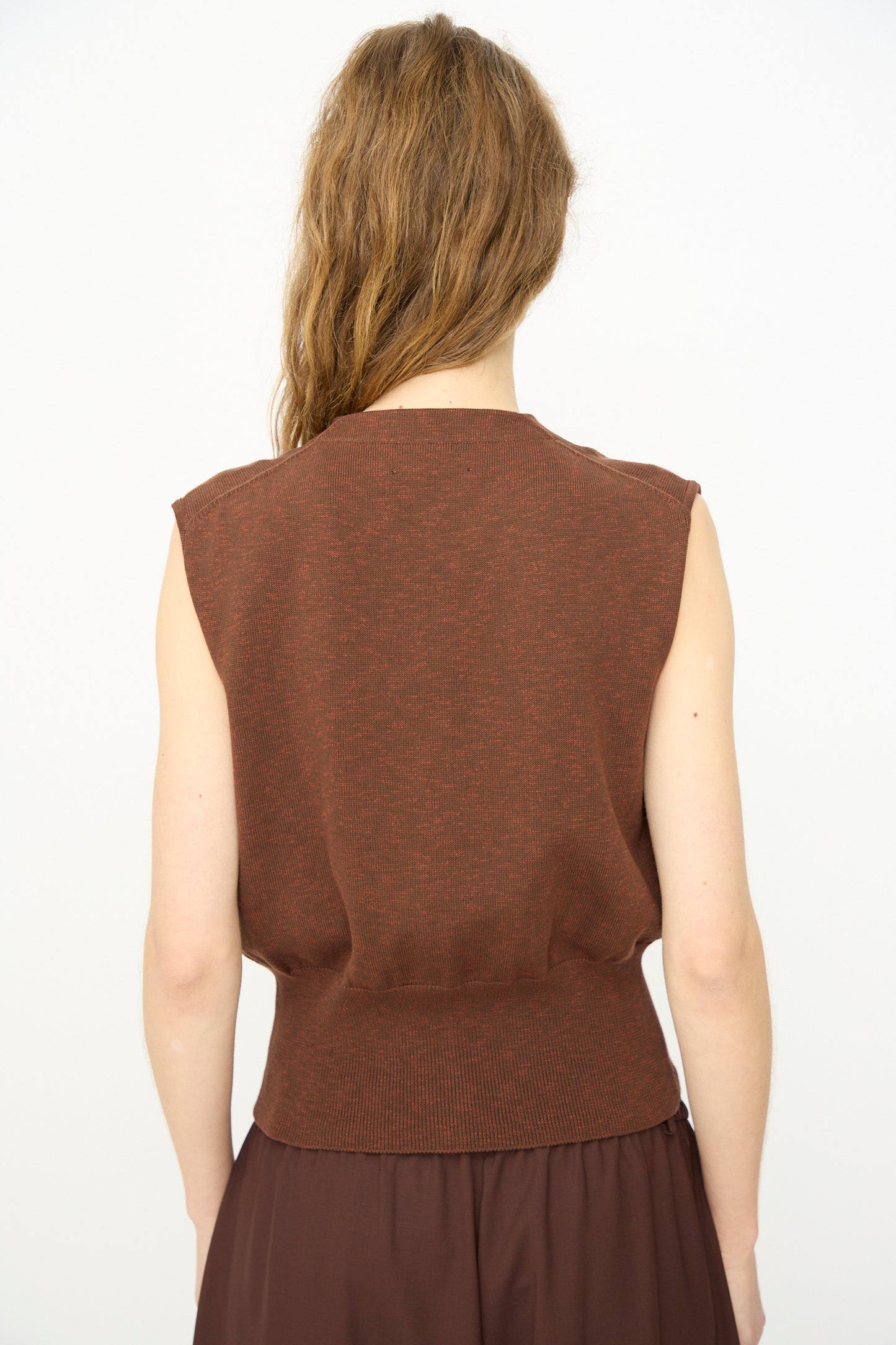 A person with long, wavy brown hair is shown from the back wearing Cordera's Flecked Cotton Waistcoat in Russet and a brown skirt made in Spain, set against a plain white background.