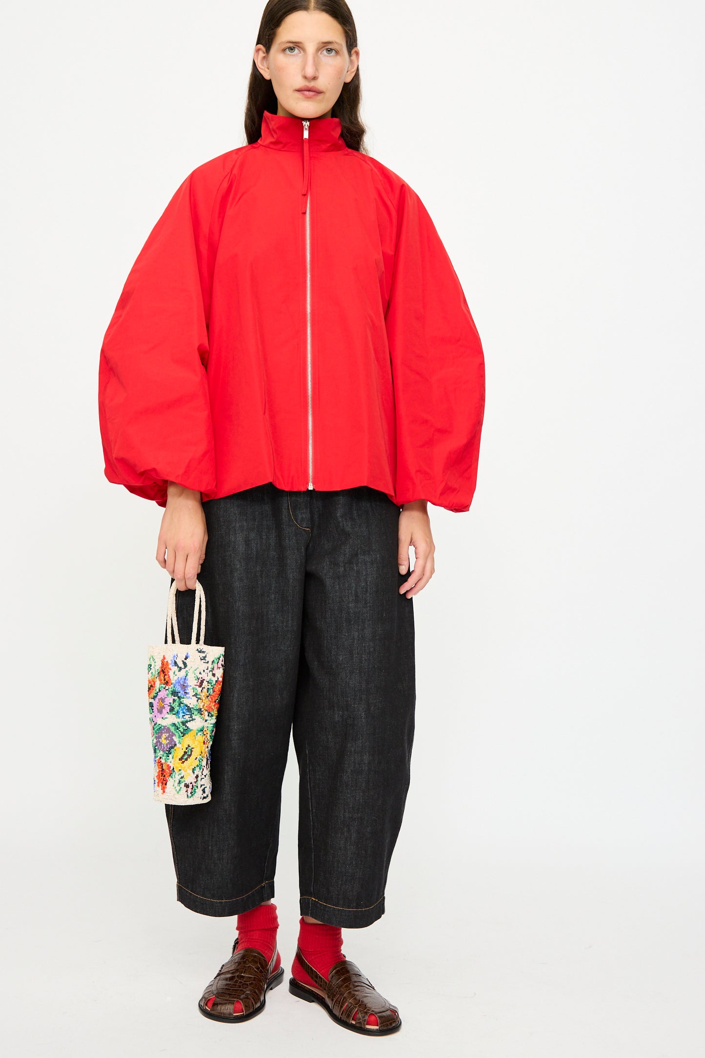 A person stands against a plain white background wearing a loose red zip-up jacket, wide-leg dark pants, red socks, brown sandals, and holding Cordera's Handmade Beaded in Crisantemo bag.