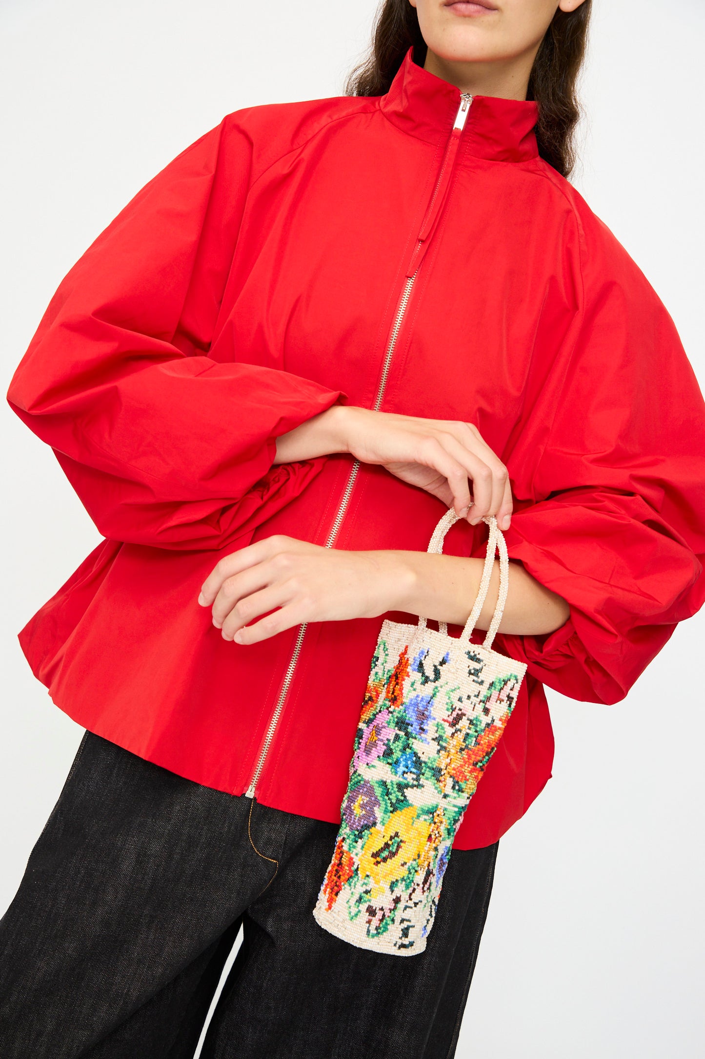 A person wearing a bright red zip-up jacket and black pants holds the Cordera Handmade Beaded in Crisantemo, a small, colorful patterned handcrafted bag with a handle.