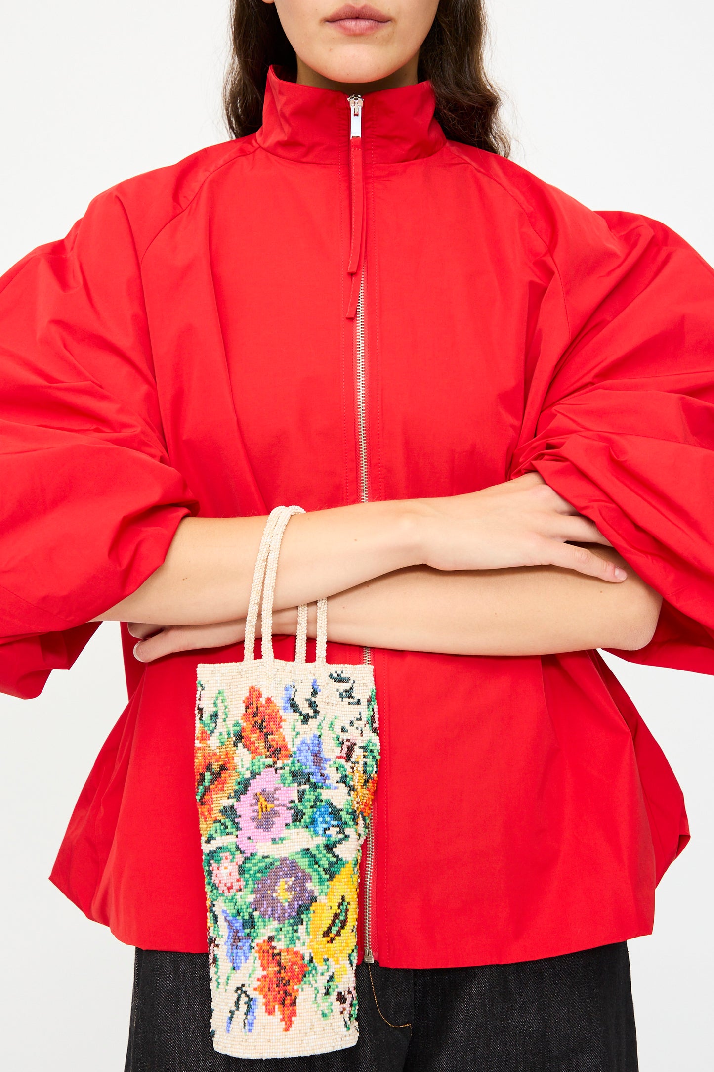 A woman in a red zip-up top with puffed sleeves stands with arms crossed, holding Cordera's Handmade Beaded in Crisantemo bag, featuring a colorful floral design.