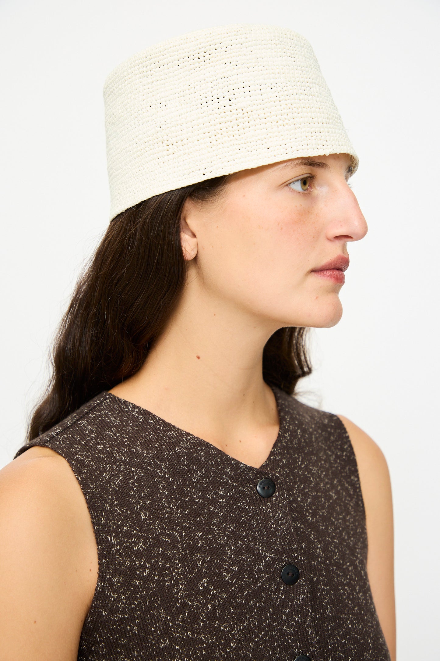 A person with long dark hair wears a sleeveless brown buttoned top and the Cordera Handmade Fez Hat in Natural, facing right against a plain background.
