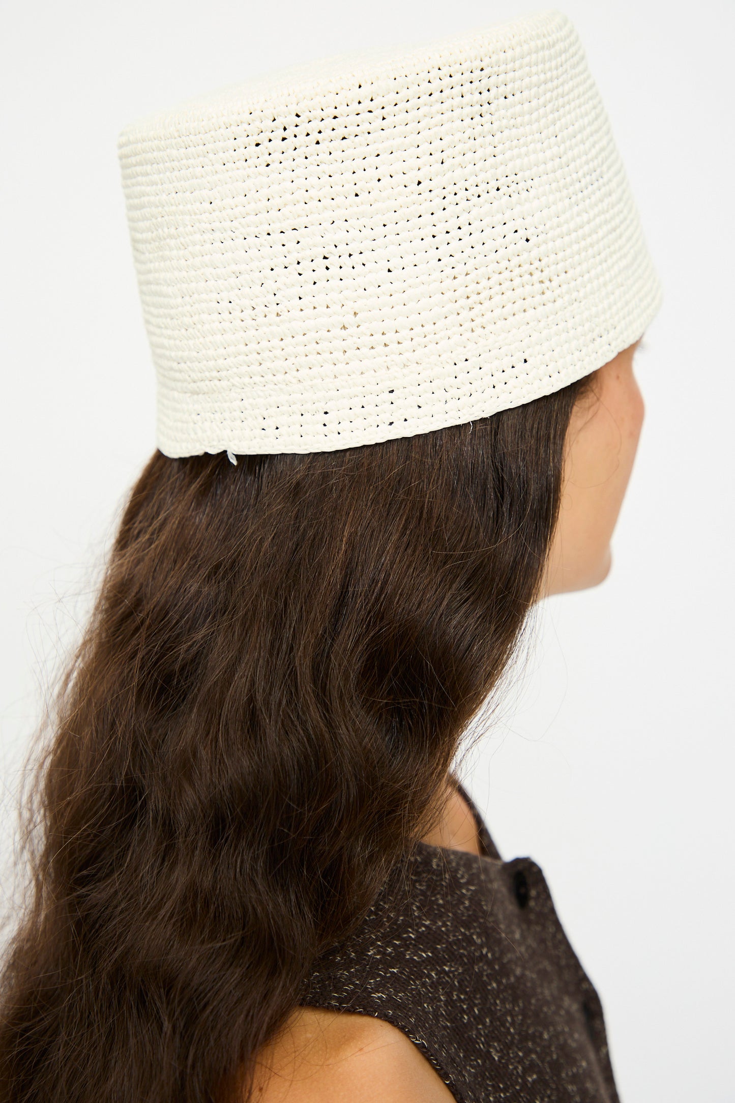 A person with long brown wavy hair wears Cordera’s Handmade Fez Hat in Natural, a textured white cylindrical hat made in Spain, paired with a sleeveless dark brown top, shown from the back and side.