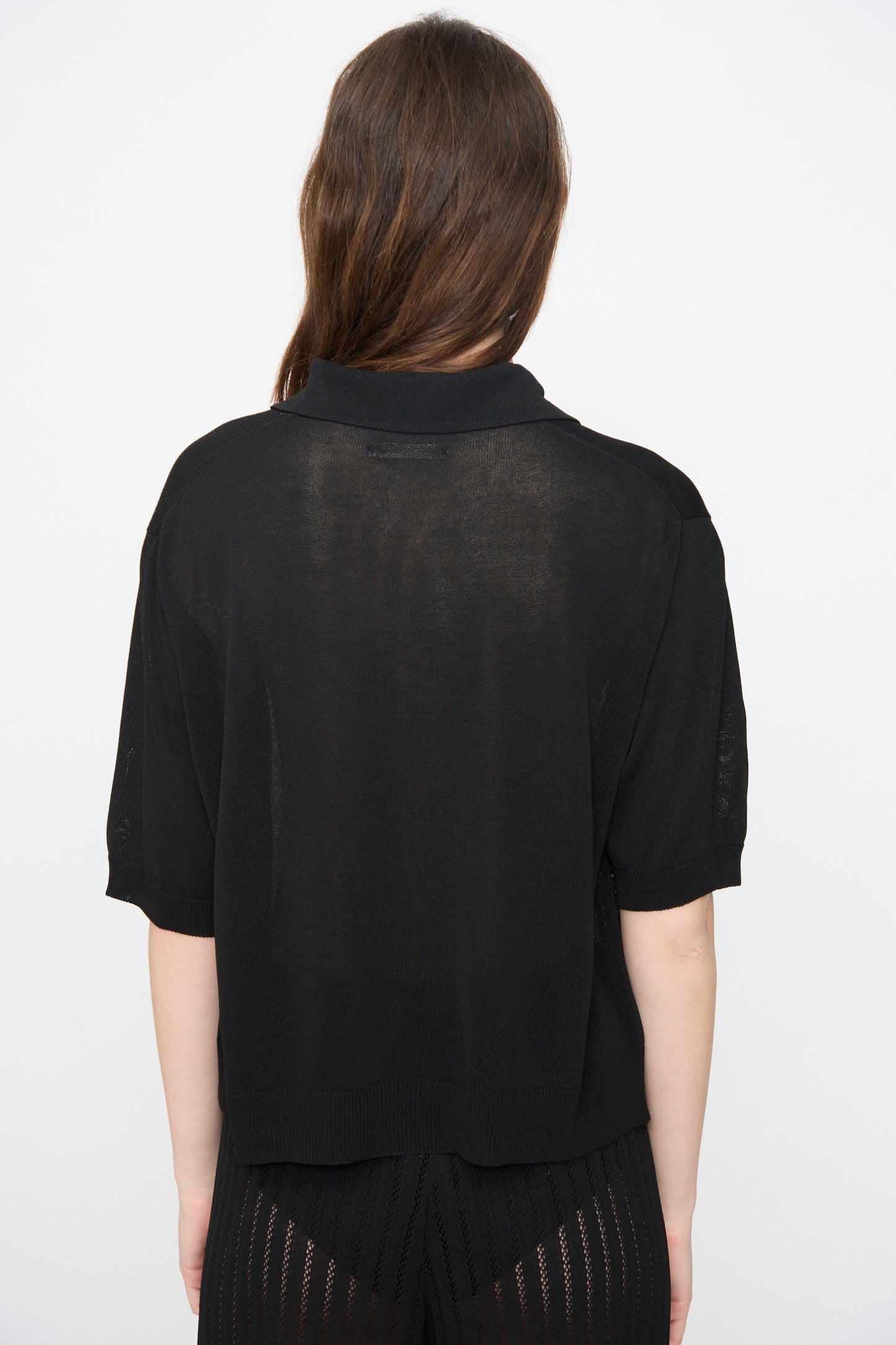 A person with long brown hair is seen from behind against a plain white background, wearing Cordera’s Lace Knit Polo in Black.