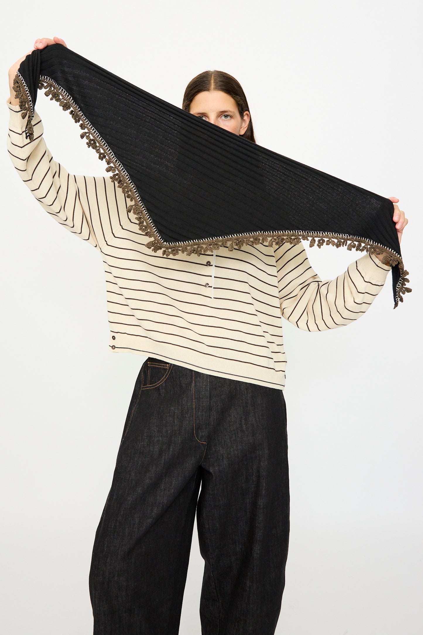 A person in a striped sweater and dark wide-leg pants holds up the Cordera Merino Wool Bandana in Black, featuring crochet lace trim and tassel edges, partially covering their face.