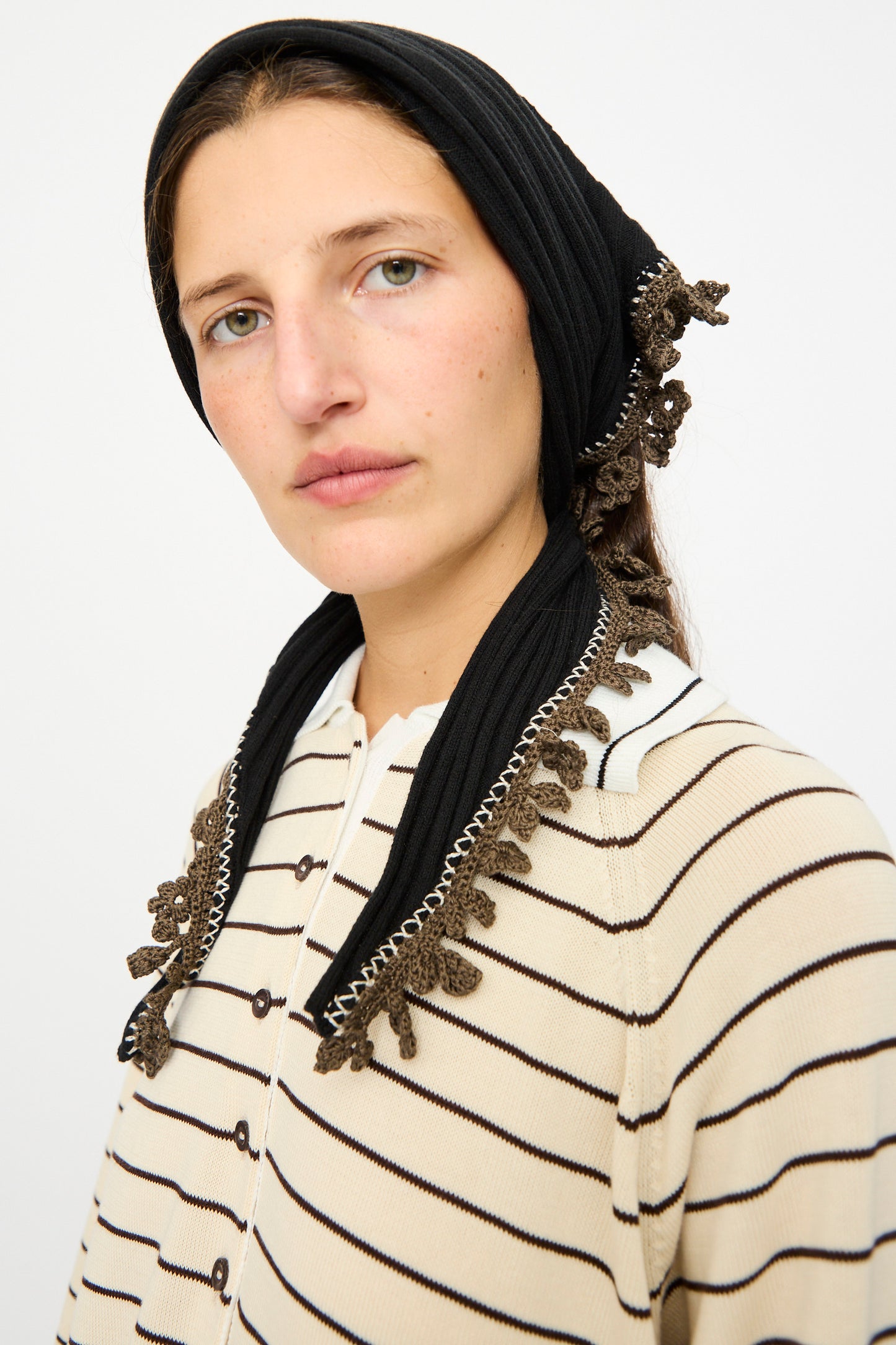 A woman wears the Cordera Merino Wool Bandana in Black, featuring brown and white crochet lace trim, paired with a beige striped sweater, looking at the camera against a plain light background.