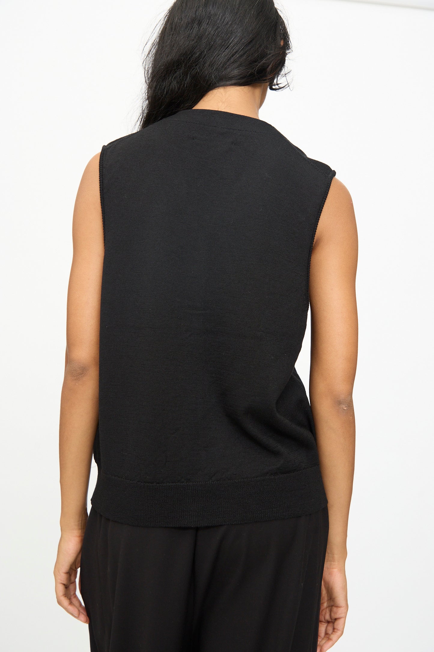 A person with long dark hair is shown from the back, wearing the Cordera Merino Wool Button Waistcoat in Black over a sleeveless black top and black pants, set against a plain white background.