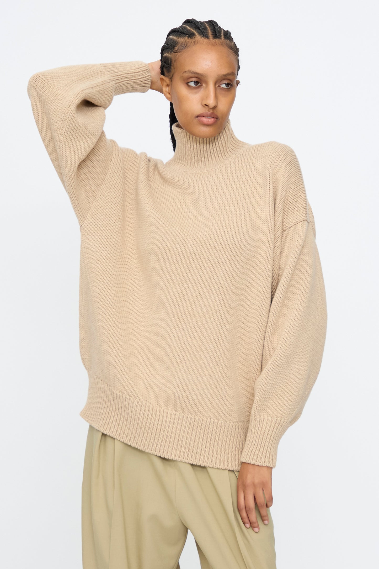 A woman poses with one arm raised behind her head, wearing the Cordera Merino Wool Chunky Turtleneck Sweater in Beige and light khaki pants, set against a plain white background.