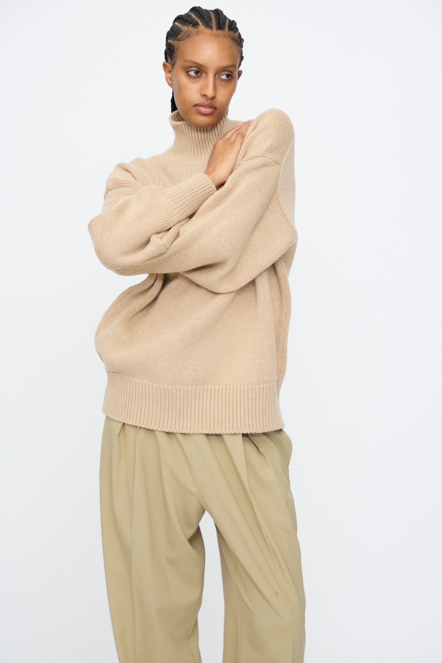 A person stands against a plain background, arms crossed, wearing Cordera's Merino Wool Chunky Turtleneck Sweater in Beige and loose khaki pants.