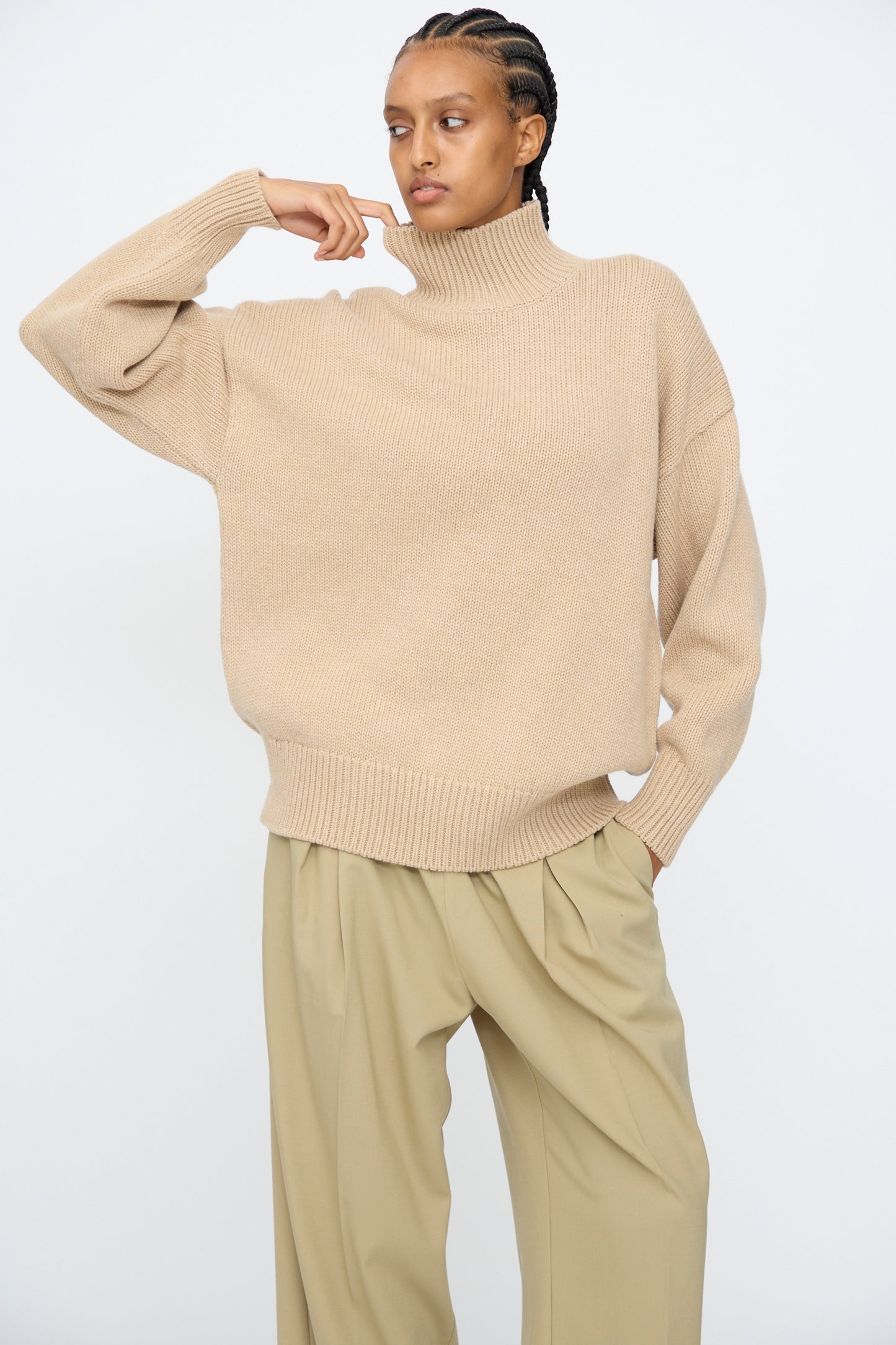 A person wearing the Cordera Merino Wool Chunky Turtleneck Sweater in Beige and wide-leg khaki pants stands against a plain white background, one hand raised near their face.