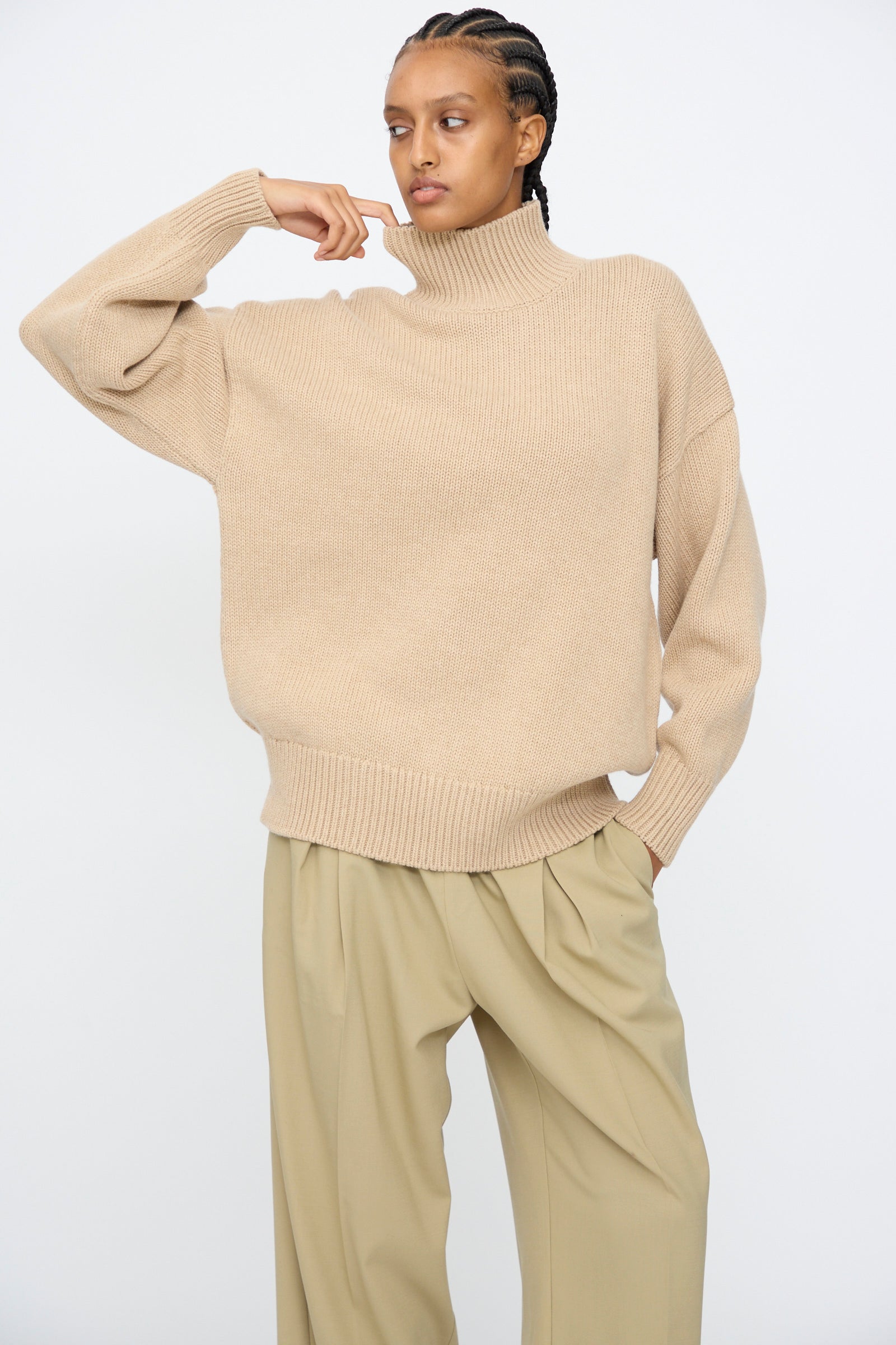 A person wearing the Cordera Merino Wool Chunky Turtleneck Sweater in Beige and wide-leg khaki pants stands against a plain white background, one hand raised near their face.