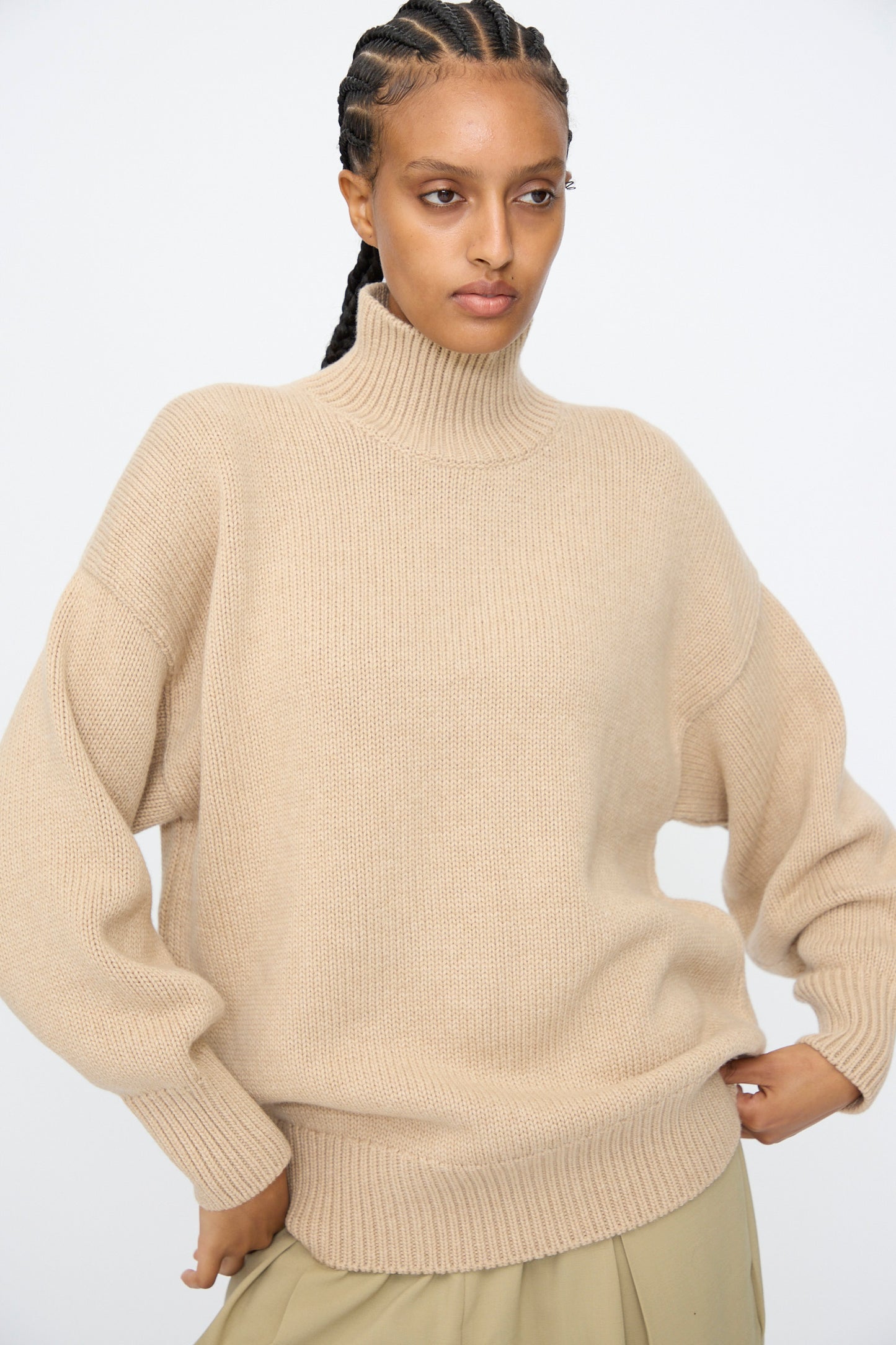 A person wears the Cordera Merino Wool Chunky Turtleneck Sweater in Beige with light tan pants, standing against a plain light background, hands on hips.
