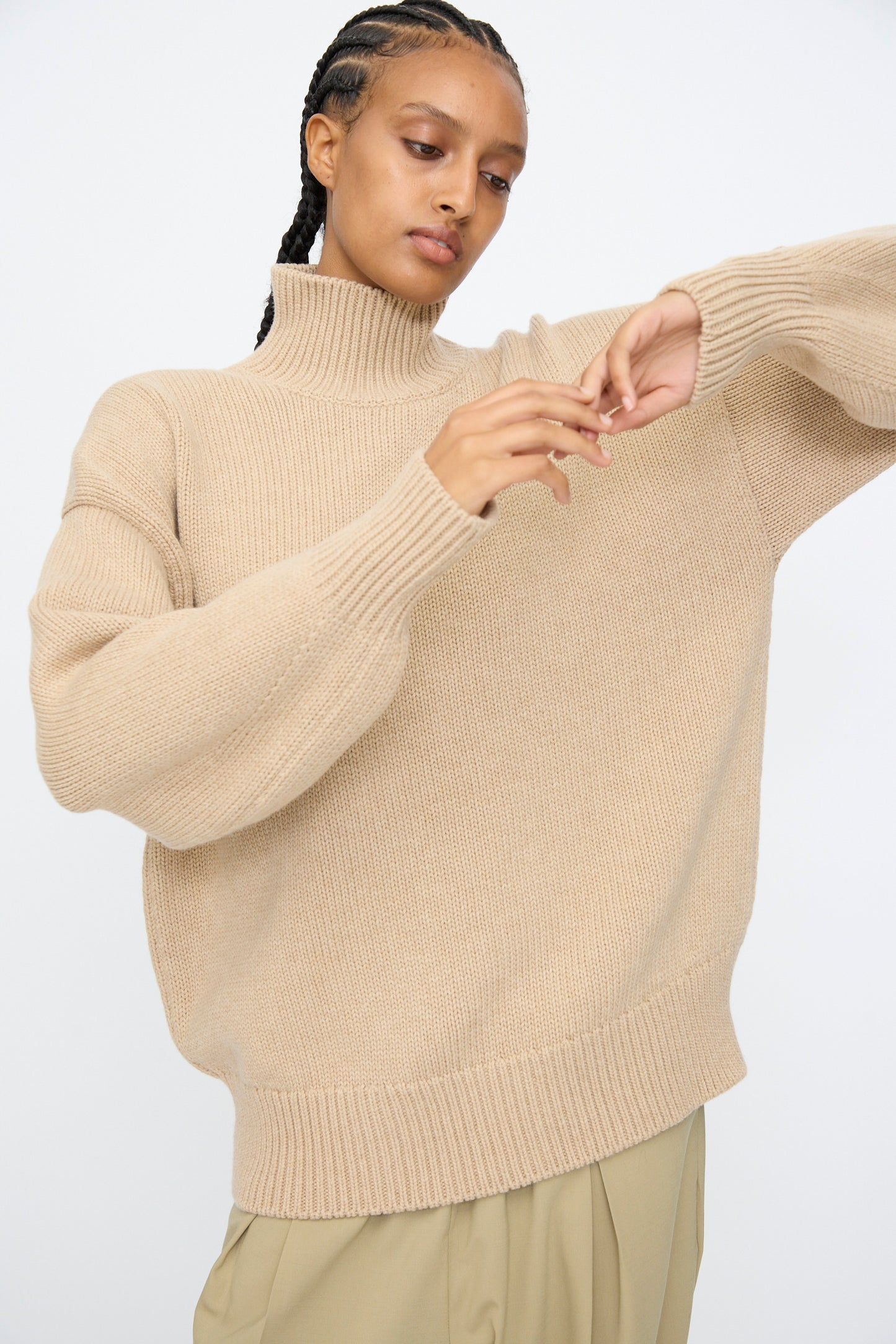 A person wears the Cordera Merino Wool Chunky Turtleneck Sweater in Beige with light pants, looking down and adjusting their hands against a plain light background.