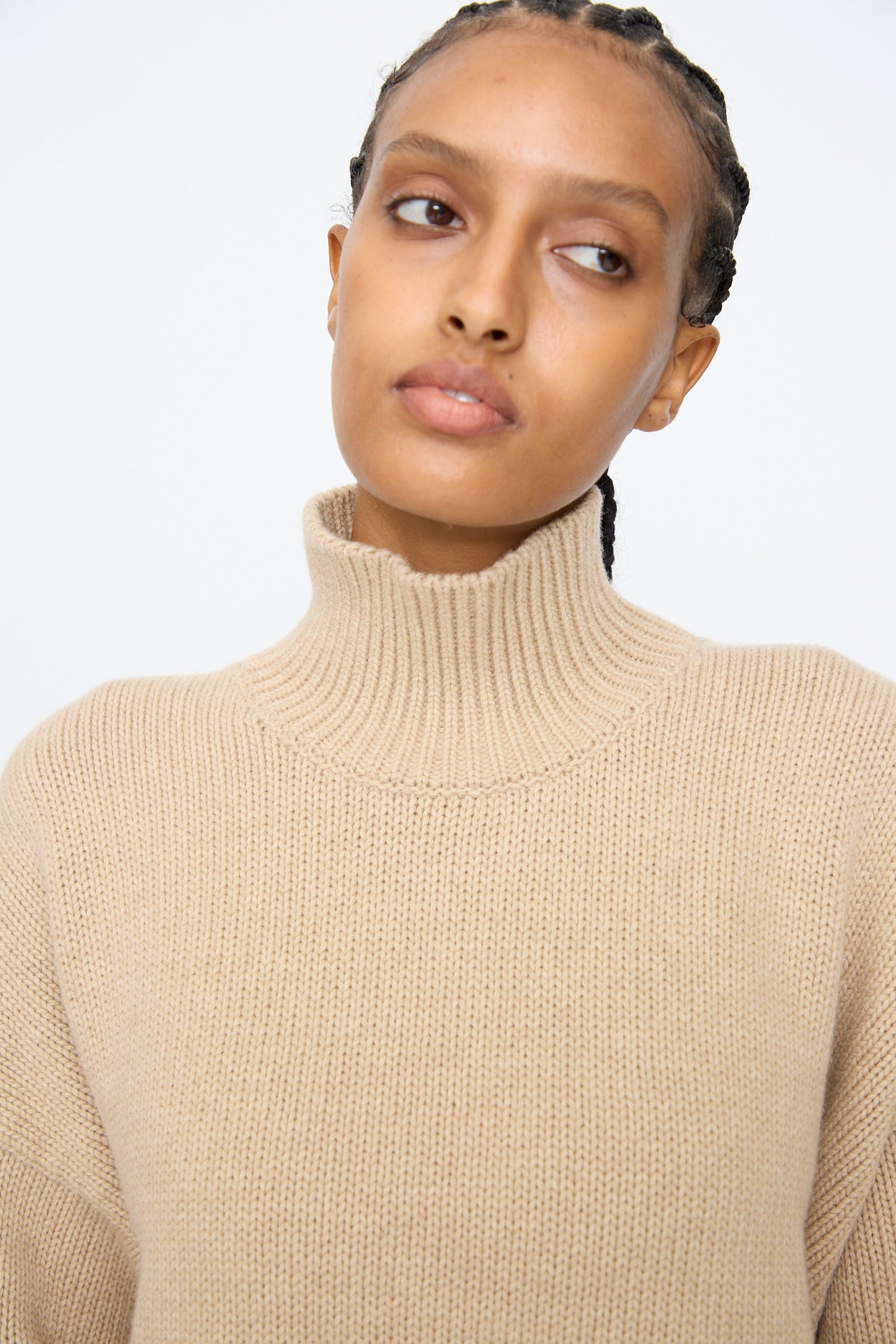 A person with braided hair, wearing the Cordera Merino Wool Chunky Turtleneck Sweater in Beige, gazes slightly to the side against a plain background.