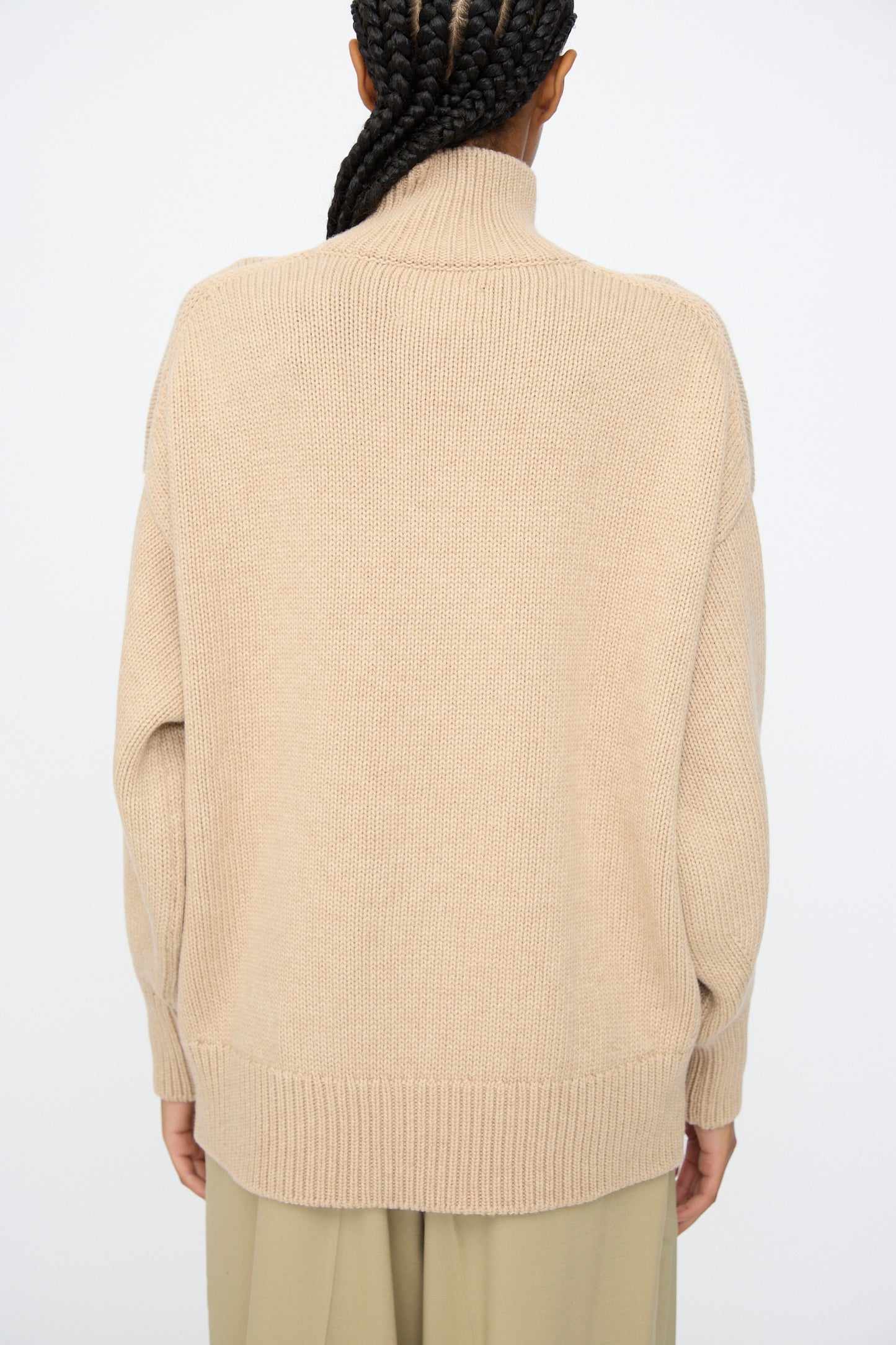 A person with braided hair is shown from the back, wearing the Cordera Merino Wool Chunky Turtleneck Sweater in Beige and light-colored pants against a plain background.