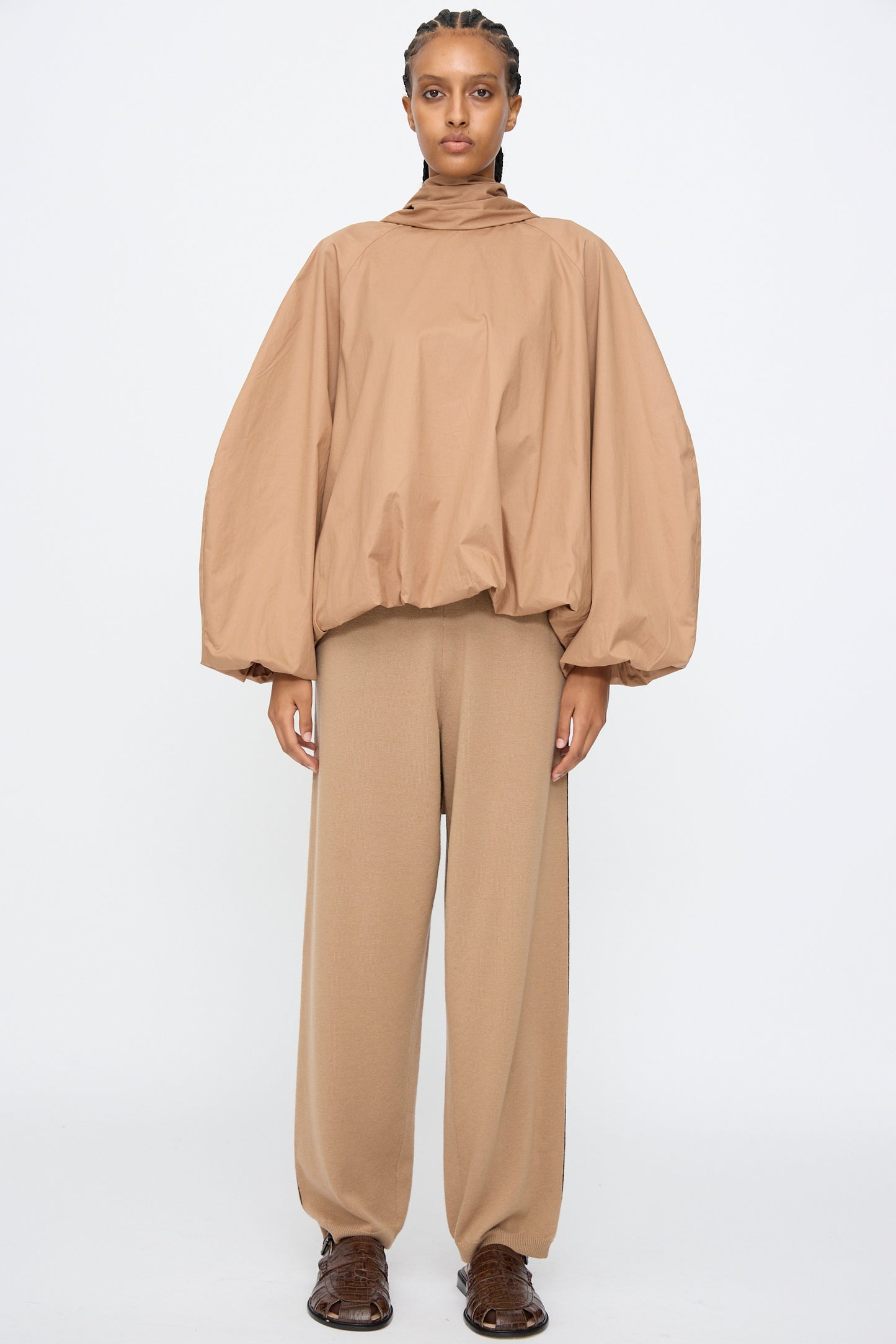 A person stands against a plain background wearing Cordera’s Merino Wool Straight Pants in Camel, paired with a matching oversized tan balloon-sleeve top and brown shoes.