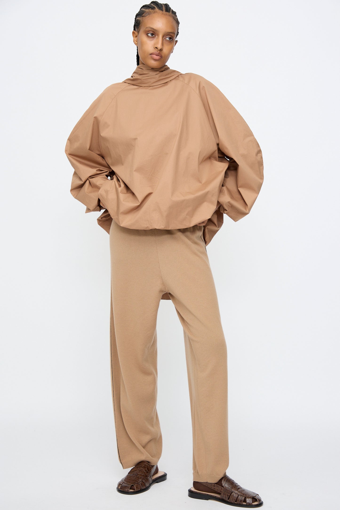 A person stands against a white background wearing Cordera’s Merino Wool Straight Pants in Camel, paired with an oversized tan top and brown flat shoes.