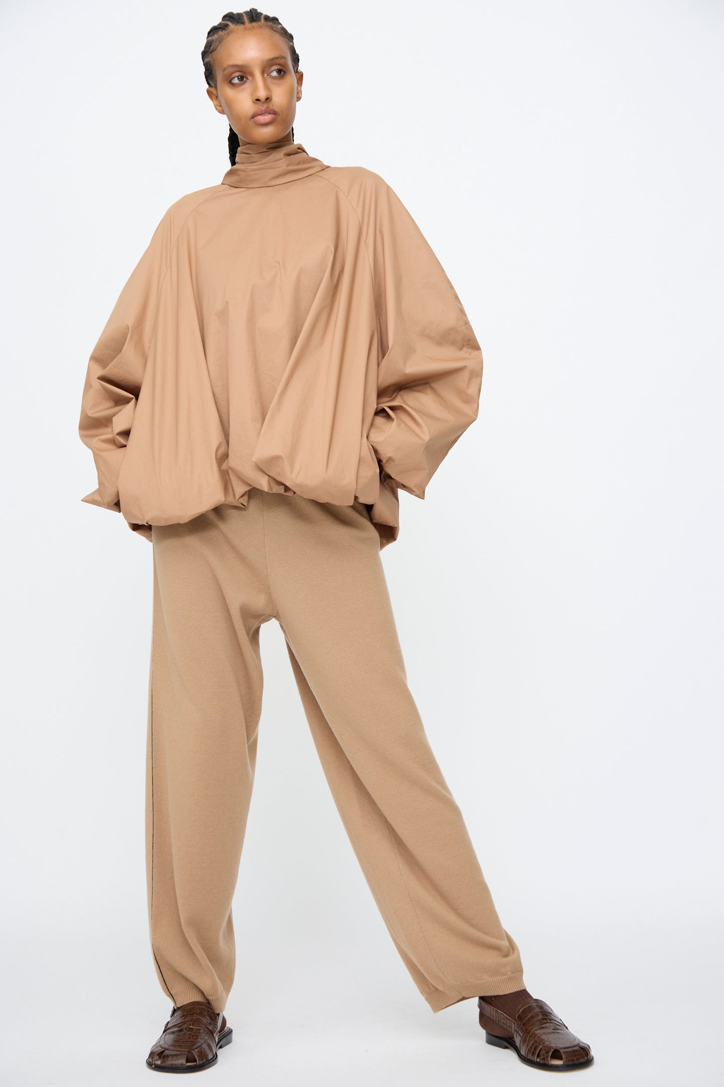 A person stands against a plain white background wearing Cordera’s Merino Wool Straight Pants in Camel, paired with an oversized tan long-sleeve top and brown shoes.