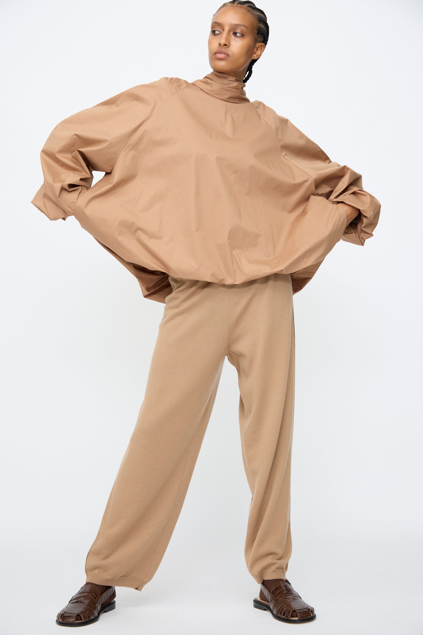 Model wears Cordera’s Merino Wool Straight Pants in Camel, styled with a loose tan long-sleeve top, standing with legs apart and hands lifting the sides of the top against a plain white background.