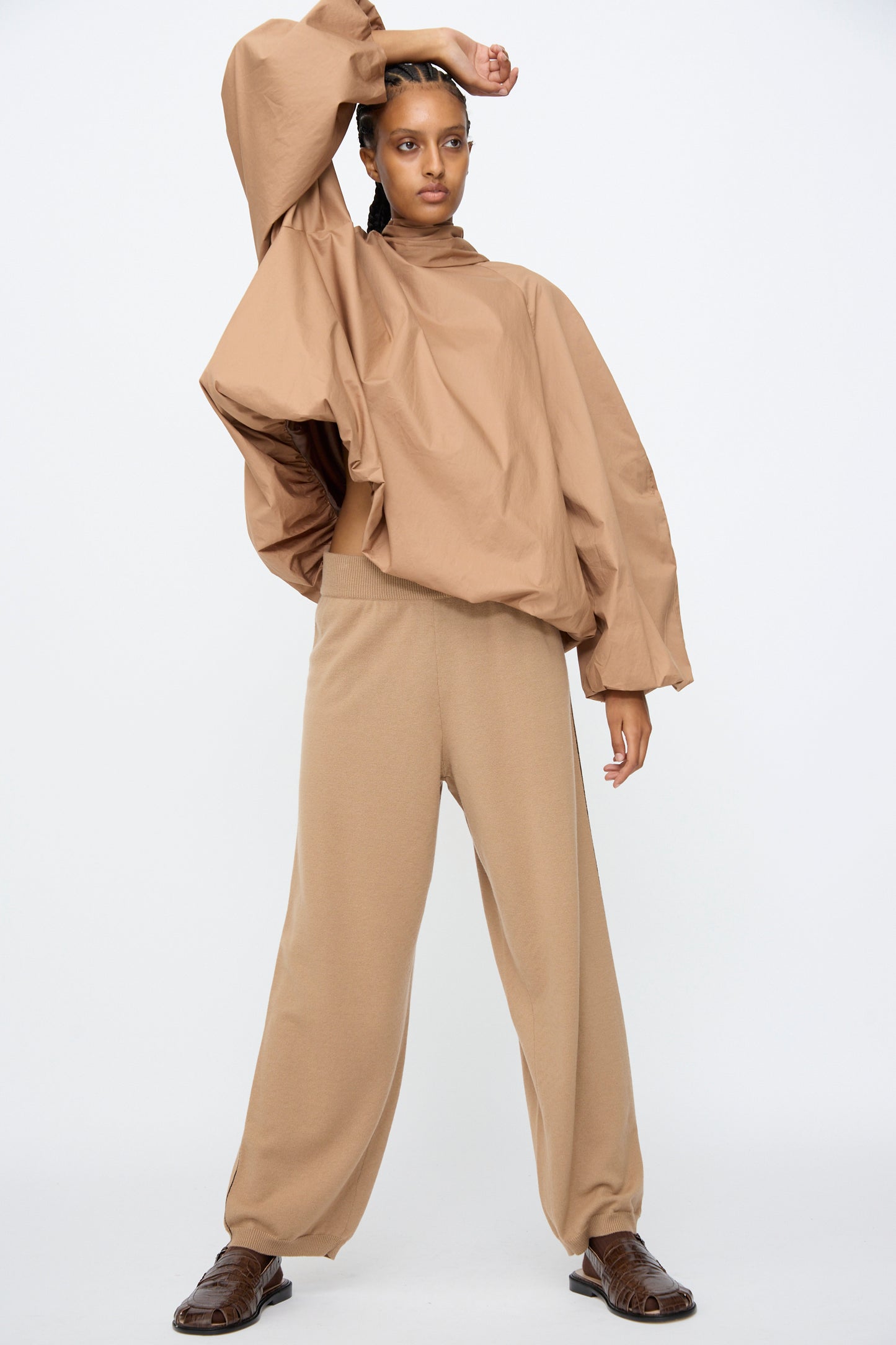 A person poses against a plain background wearing a loose tan long-sleeve top, brown shoes, and Cordera Merino Wool Straight Pants in Camel with an oversized fit and wide sleeves.