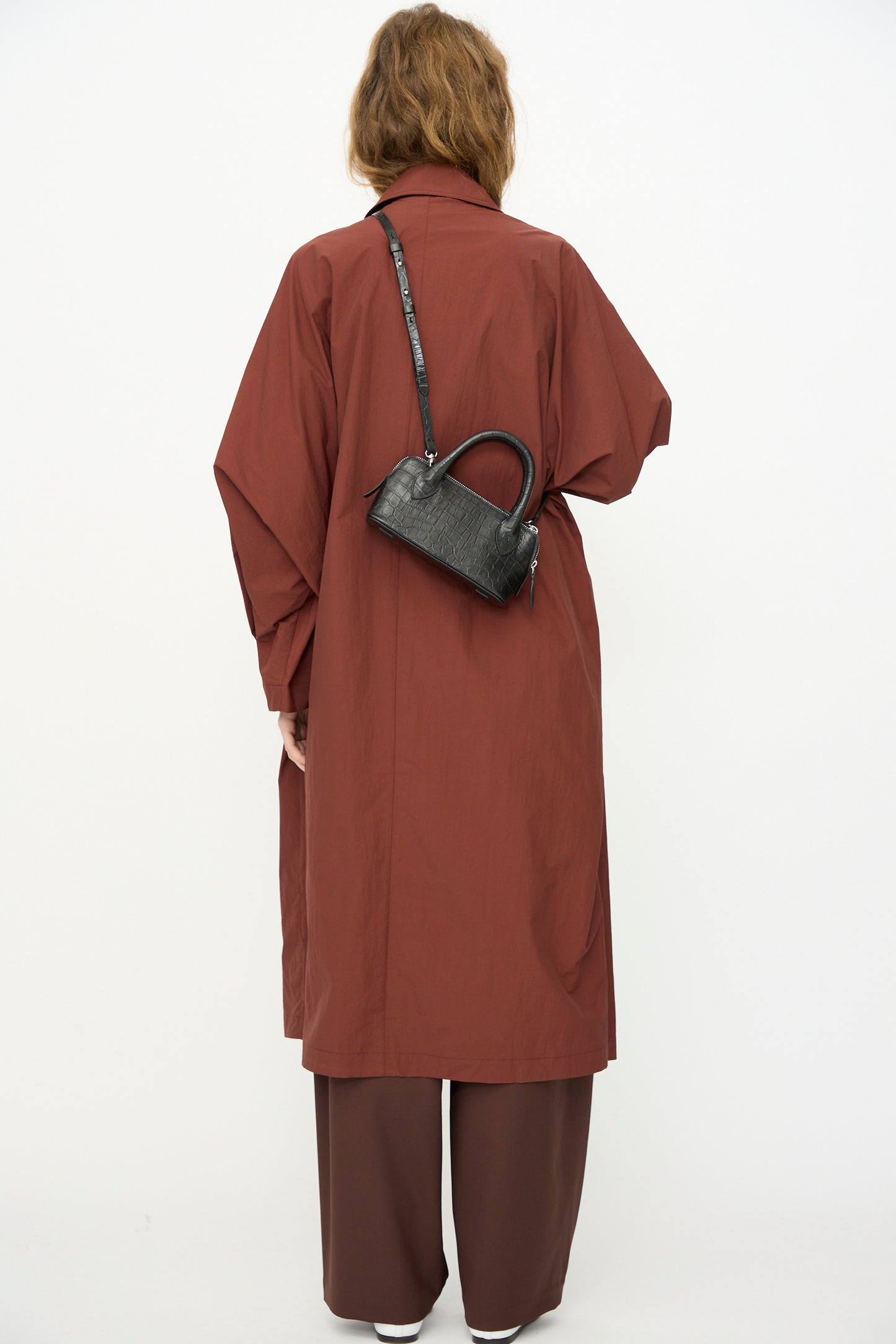 Person facing away in a long brown coat and matching pants, with the Cordera Mini Bag Crocodile in Black worn crossbody, stands against a plain white background.