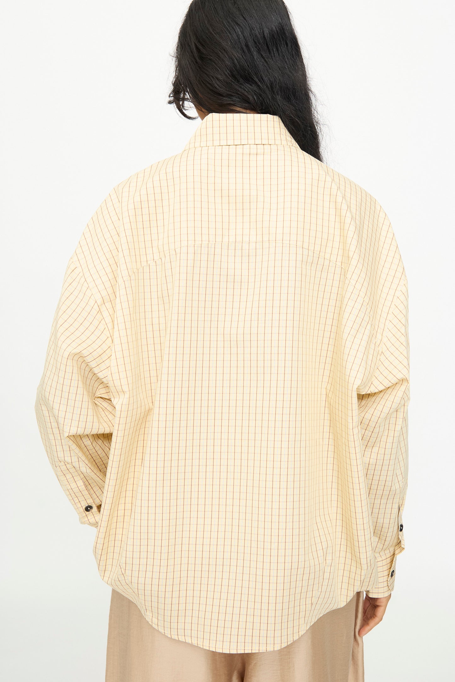 A person with long dark hair, viewed from the back, wears the Cordera Oversized Shirt in Checked—an oversized beige button-up featuring thin vertical stripes and an Italian collar.
