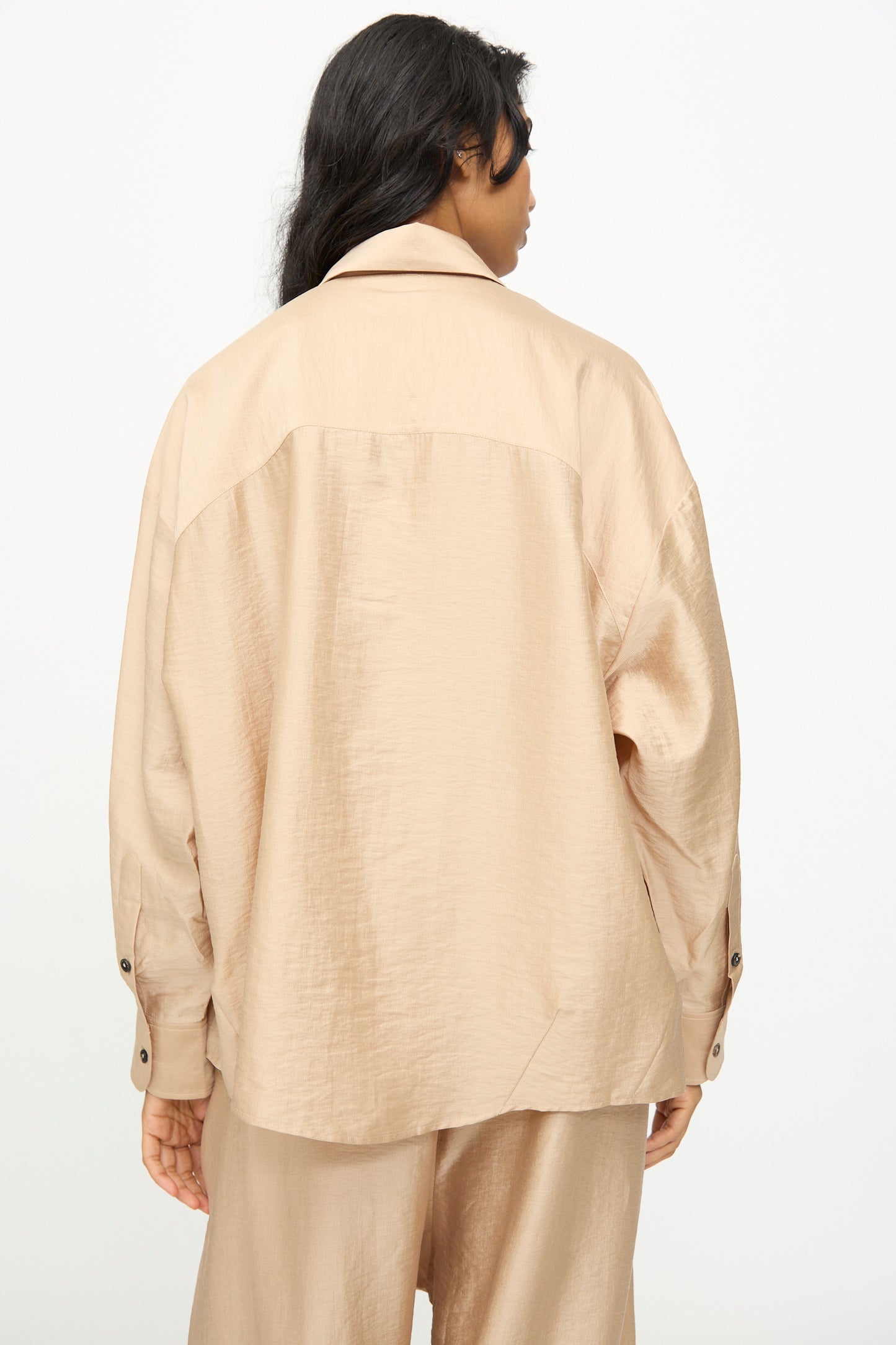 A person facing away, dressed in the Cordera Oversized Shirt in Beige—an Italian-collared, long-sleeve button-up crafted from a comfortable lyocell blend—with matching pants, set against a plain white background.
