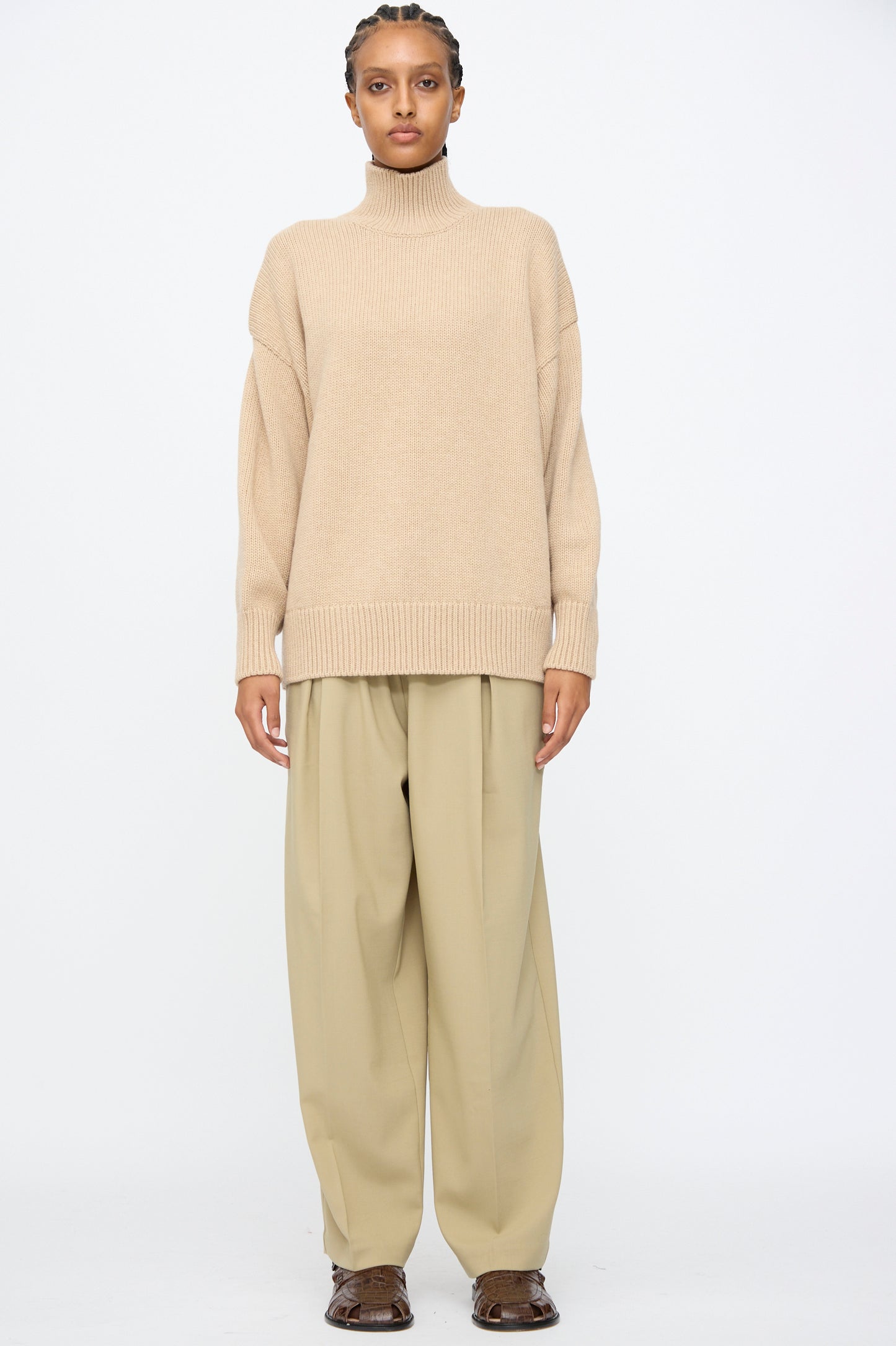 A person stands against a plain background, wearing a beige turtleneck sweater, Cordera Pleated Relaxed Pants in Olive Grey with an elastic waistband, and brown shoes.