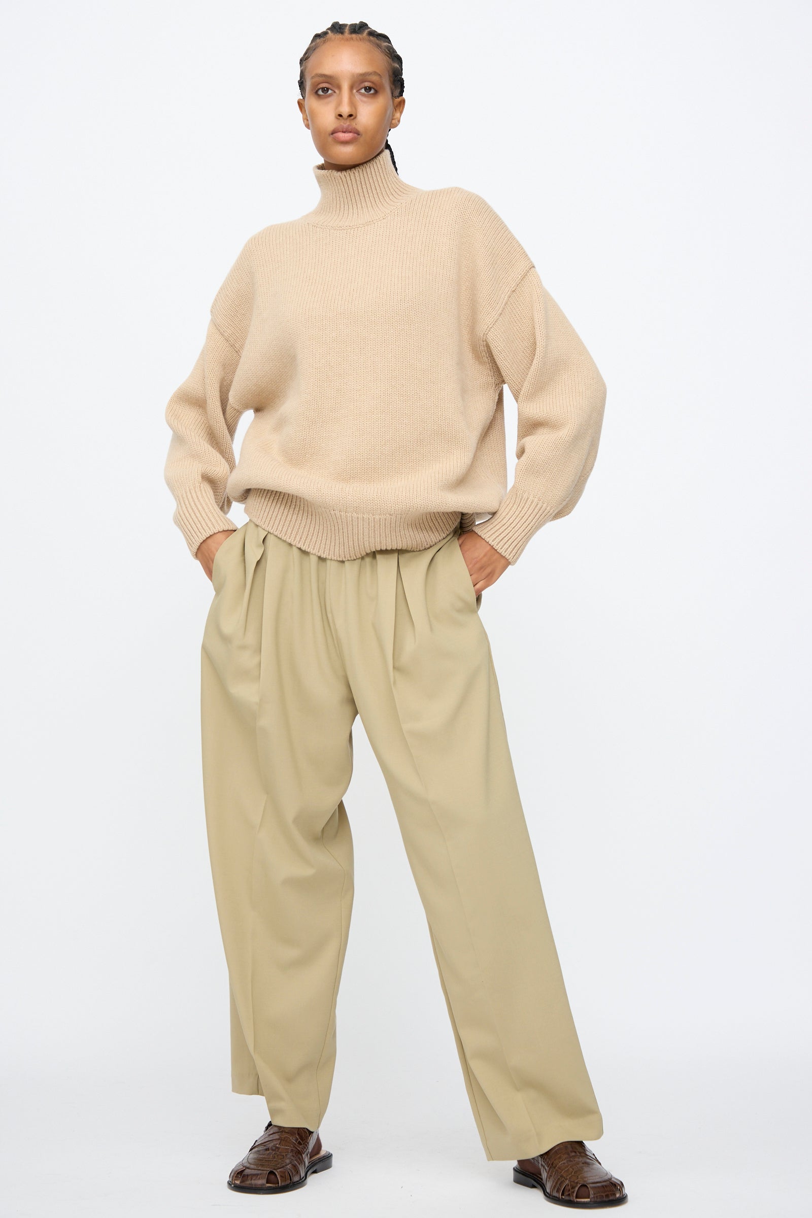 A person wearing a beige turtleneck sweater, Cordera Pleated Relaxed Pants in Olive Grey, and brown shoes stands against a plain white background.