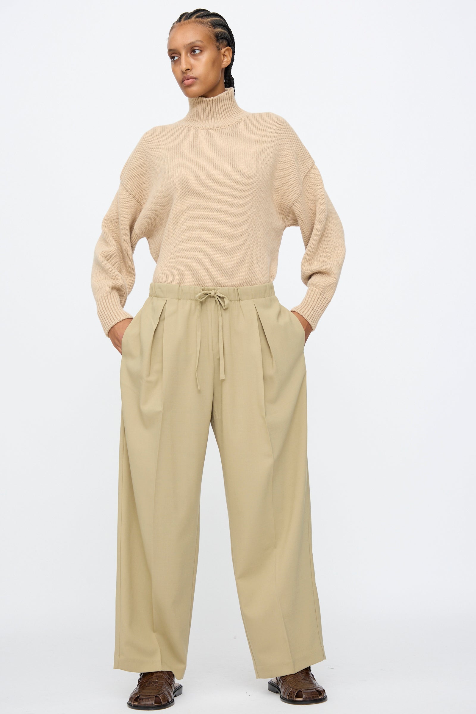 A person wearing a beige turtleneck sweater, Cordera Pleated Relaxed Pants in Olive Grey, and brown shoes stands against a plain white background.