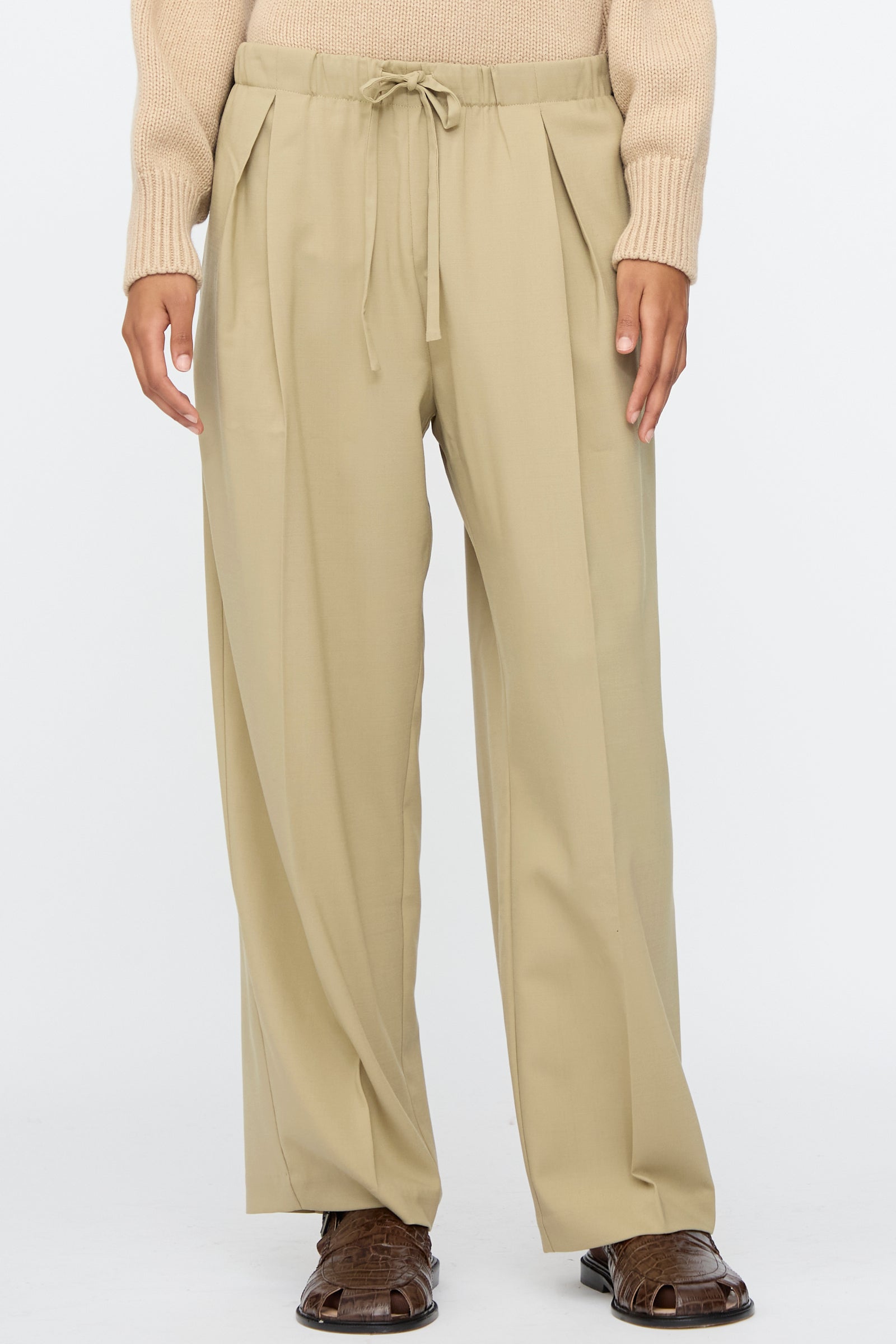 Person wearing Cordera Pleated Relaxed Pants in Olive Grey with an elastic waistband, paired with a light tan sweater and brown open-toe sandals, standing against a plain light background.