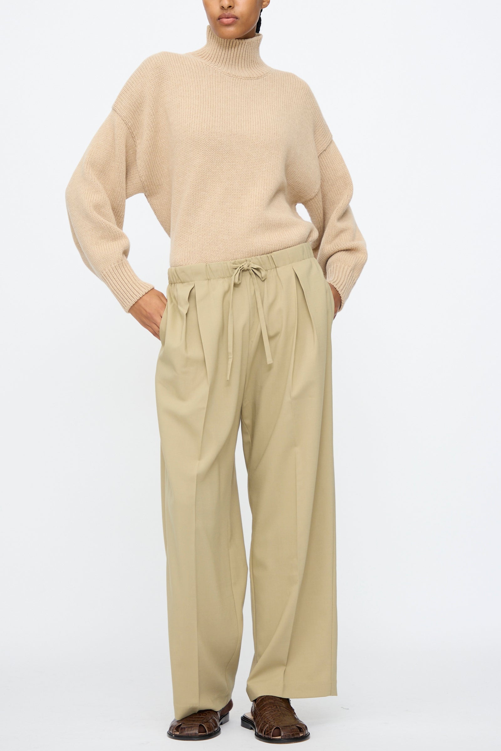 A person stands against a plain white background wearing a beige turtleneck sweater, Cordera Pleated Relaxed Pants in Olive Grey with an elastic waistband, and brown shoes.