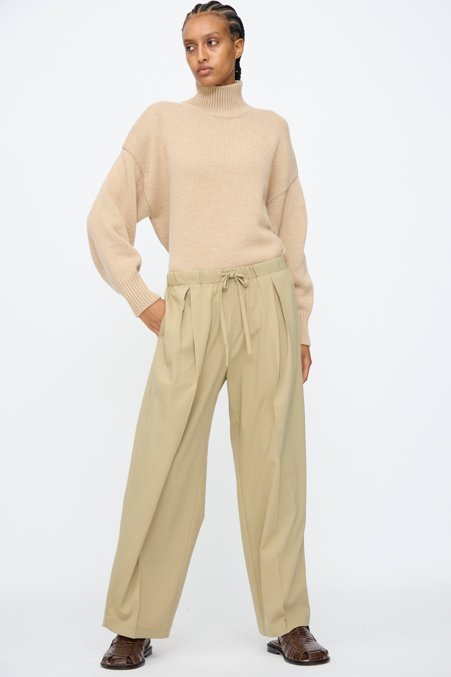 A person stands against a plain background, wearing a beige turtleneck sweater and Cordera’s Pleated Relaxed Pants in Olive Grey, featuring lightweight wool fabric and an elastic waistband, paired with brown shoes.