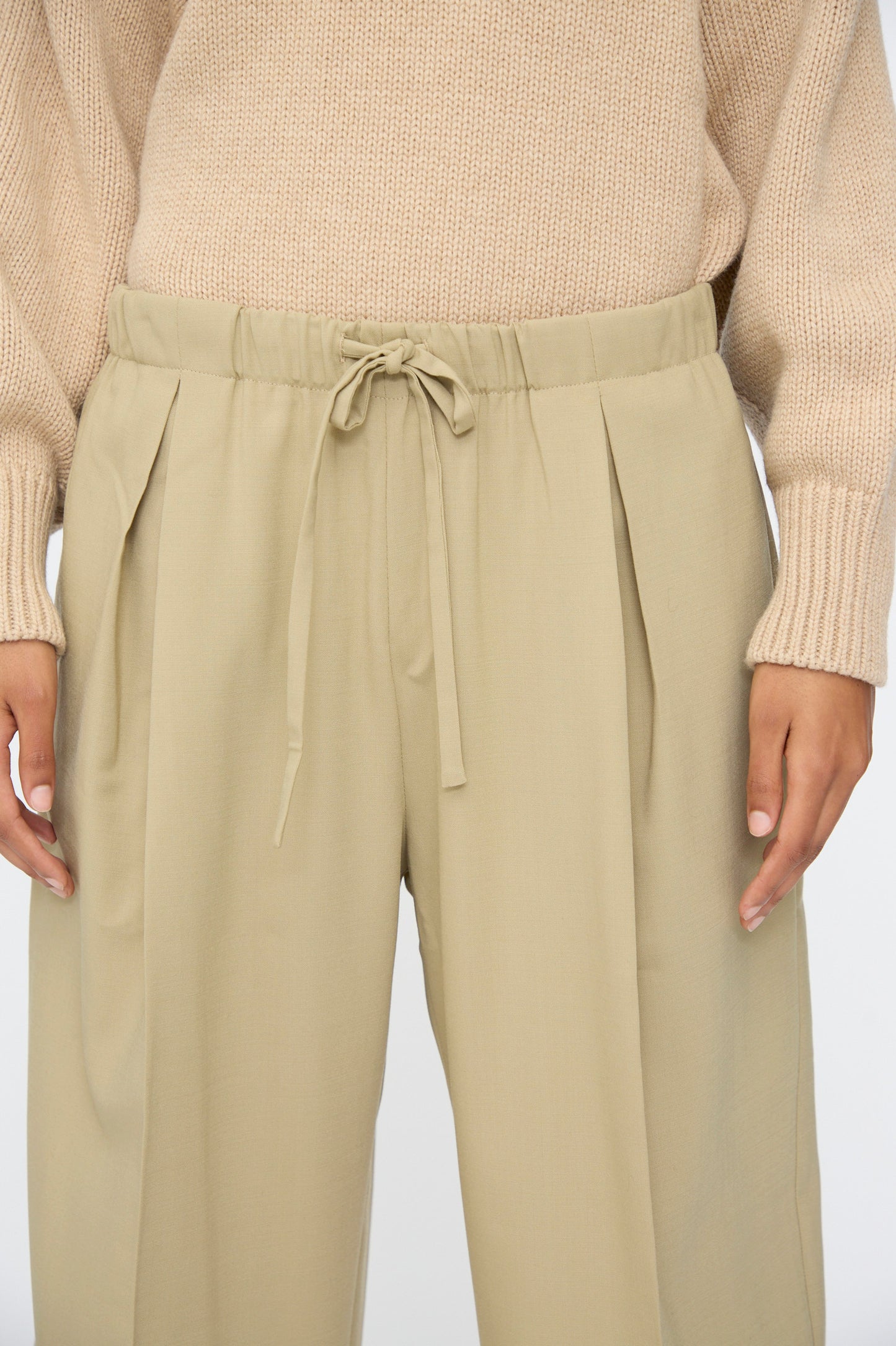 A person is shown from mid-torso to thighs, wearing a beige knit sweater and Cordera's Pleated Relaxed Pants in Olive Grey with an elastic waistband.