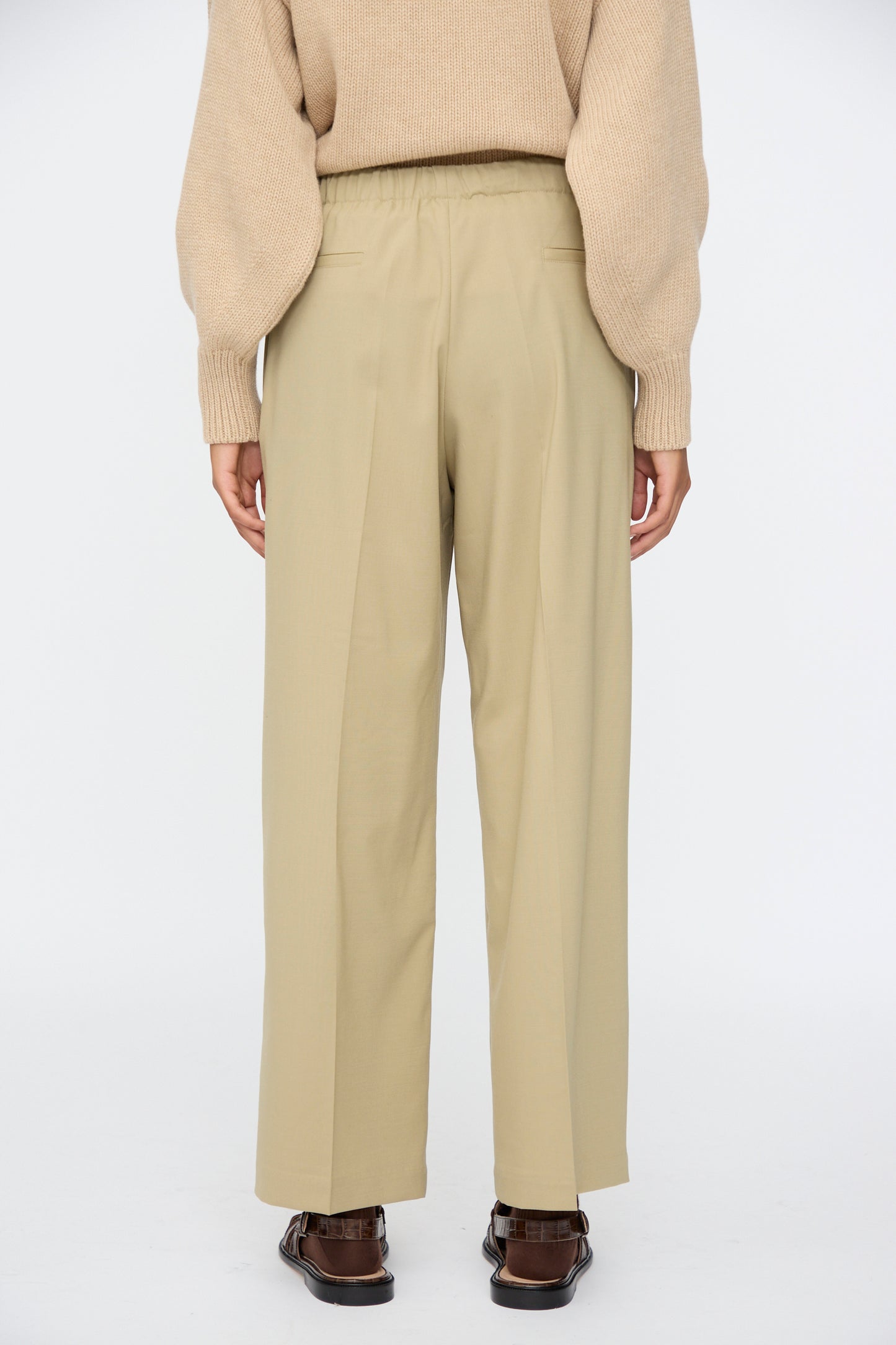 A person stands facing away against a white background, wearing a beige sweater, Cordera Pleated Relaxed Pants in Olive Grey, and brown shoes.
