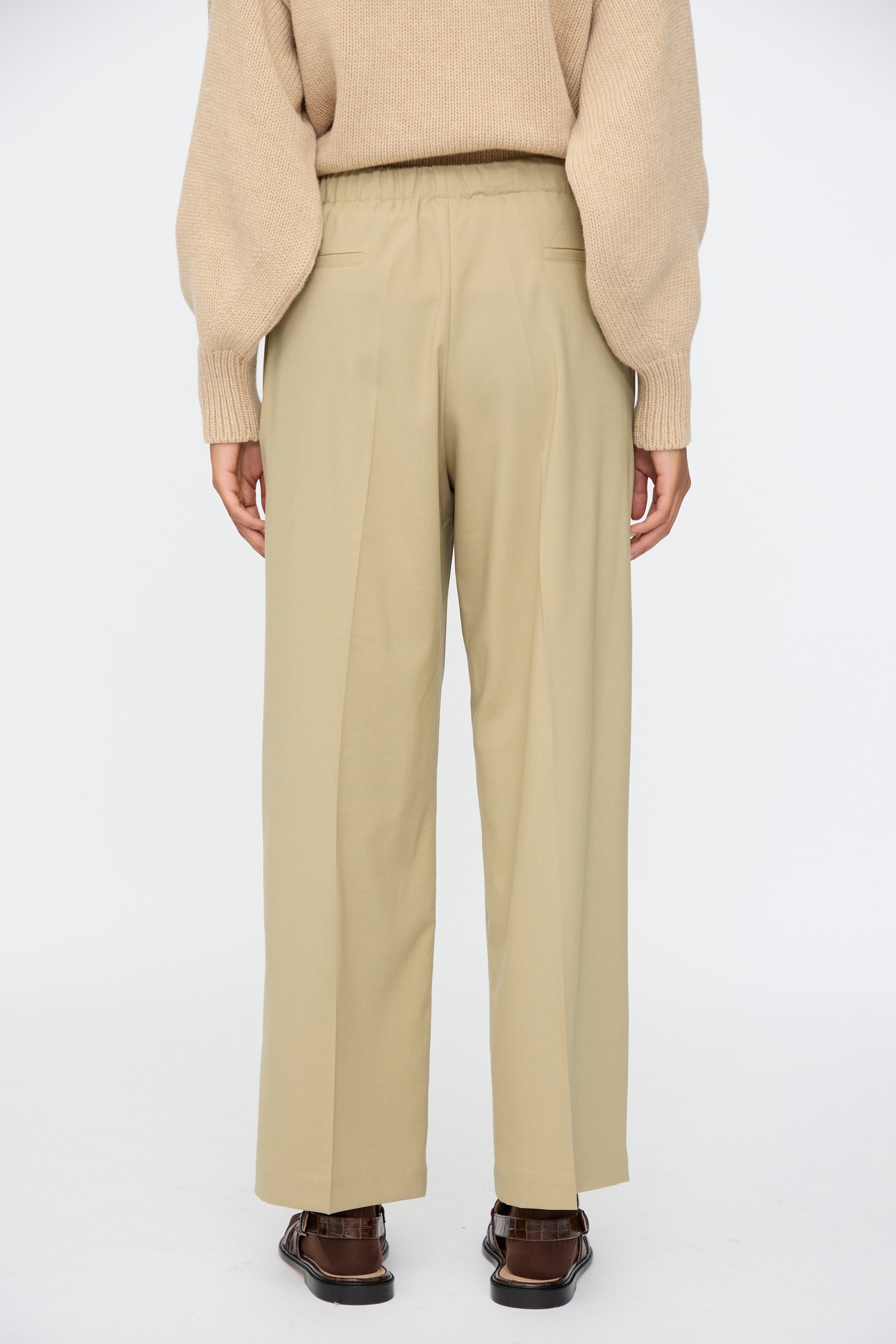 A person stands facing away against a white background, wearing a beige sweater, Cordera Pleated Relaxed Pants in Olive Grey, and brown shoes.