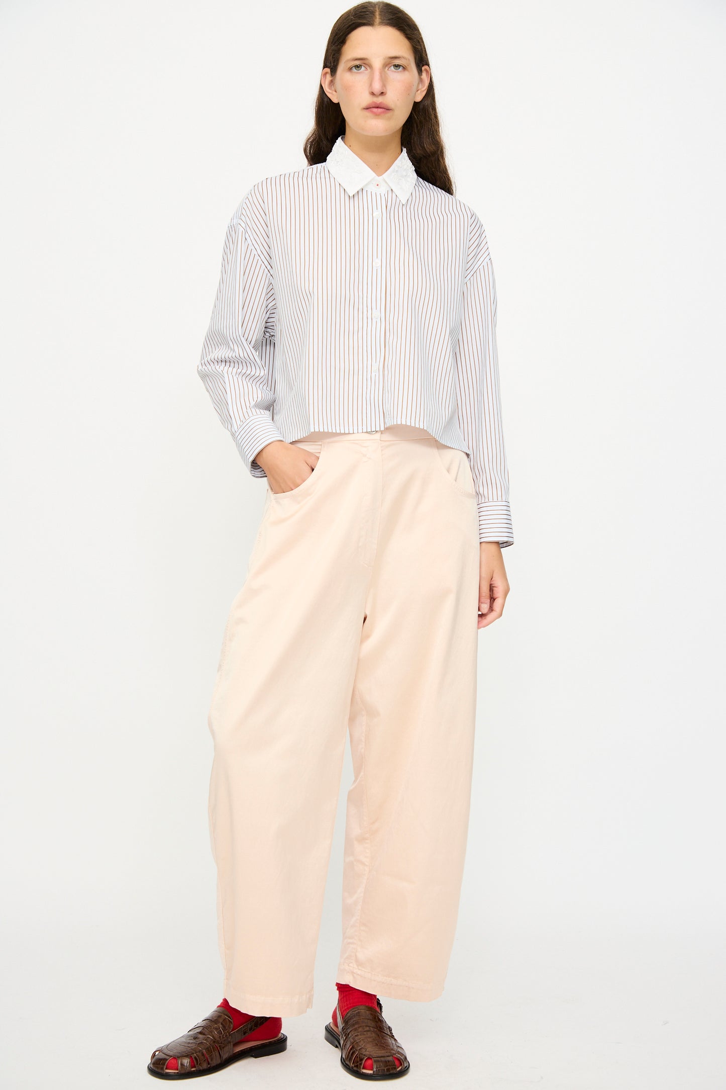 A person stands against a white background wearing a striped button-up shirt, Cordera Satin Curved Pant in Peach with an elasticated waist, brown sandals, and red socks.