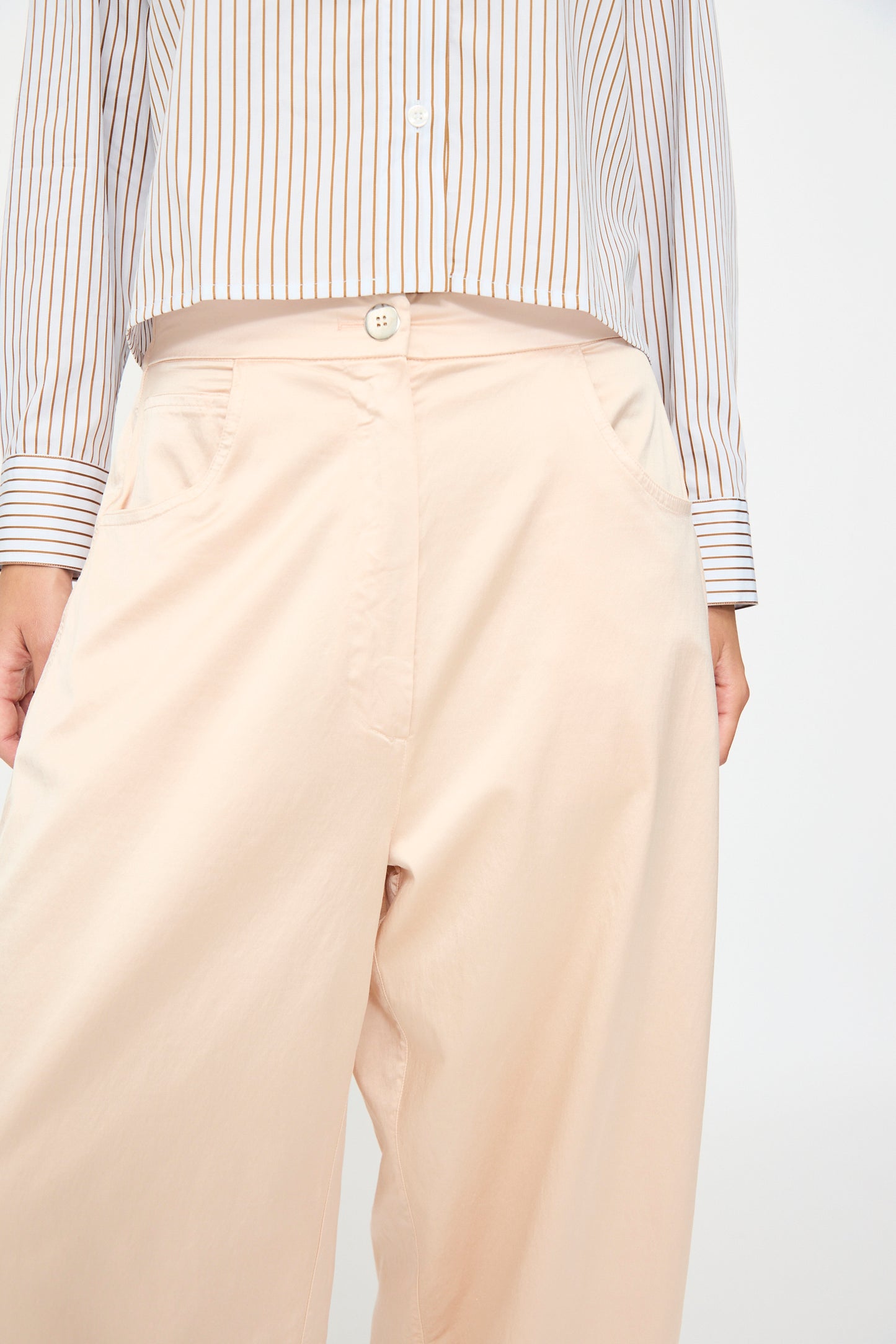 A person wears the Cordera Satin Curved Pant in Peach, featuring an elasticated waist, paired with a white button-up shirt with vertical brown stripes, shown from mid-torso to mid-thigh.