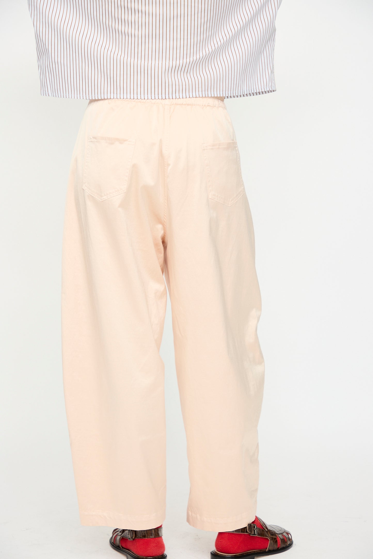 A person wearing Cordera's Satin Curved Pant in Peach, featuring an elasticated waist, styled with a blue-and-white striped shirt and red sandals, shown from the back against a plain white background.