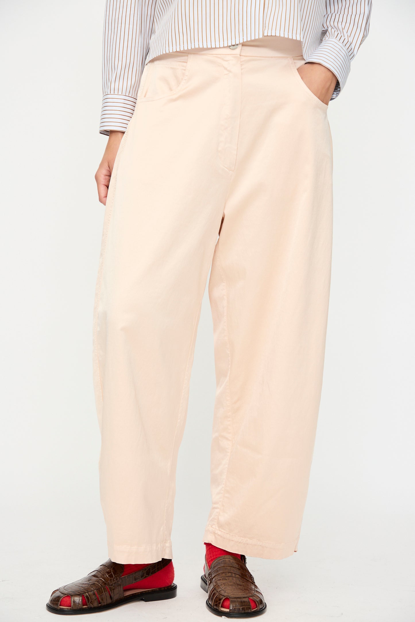 A person wearing the Cordera Satin Curved Pant in Peach with an elasticated waist, a striped shirt, red socks, and brown sandals stands against a plain background, hands tucked in their pockets.