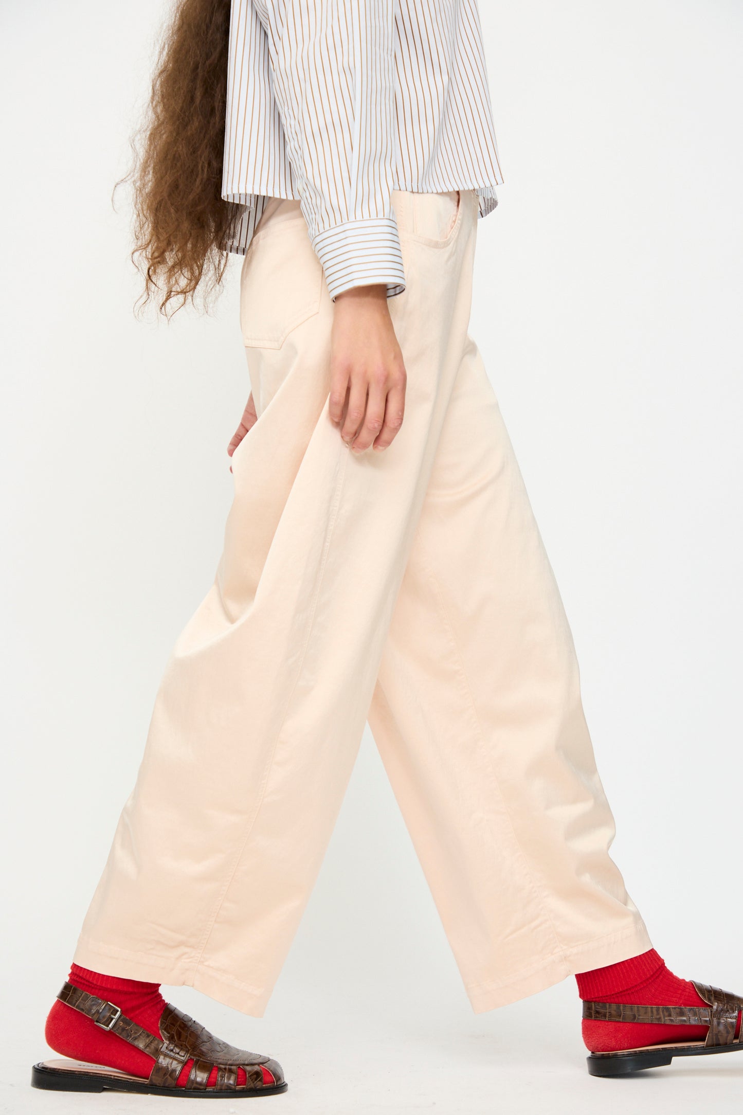Model wears Cordera’s Satin Curved Pant in Peach with a striped long-sleeve shirt, red socks, and brown sandals, walking sideways against a plain background.