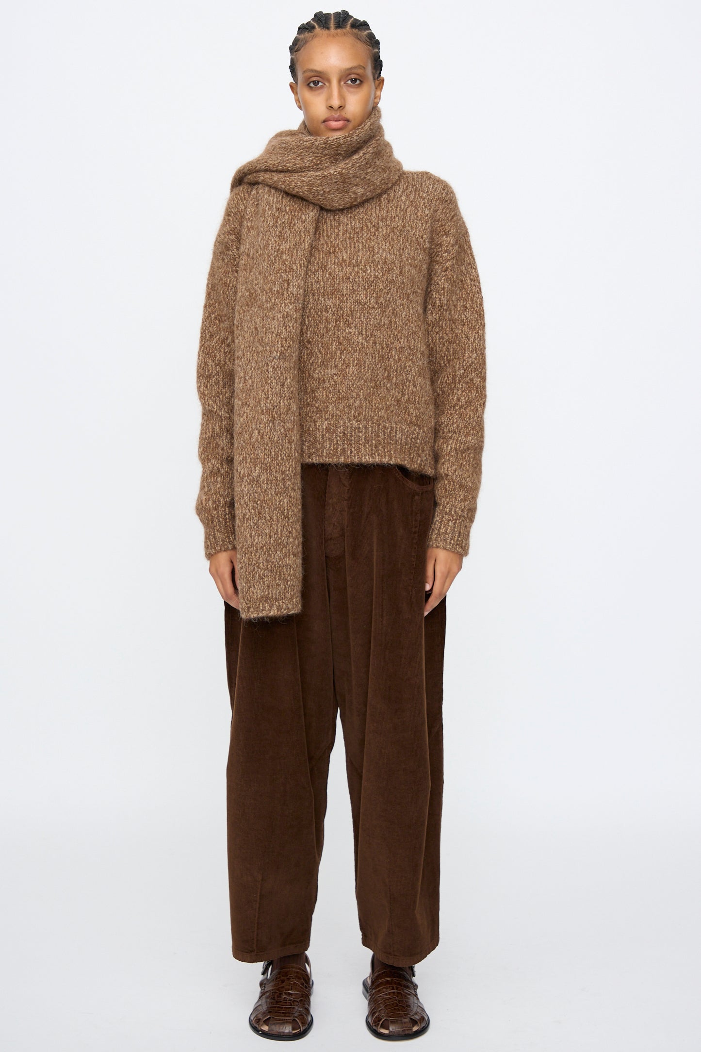 A person stands against a plain background, hands by their sides, wearing loose brown pants, a brown knit sweater, brown shoes, and the Cordera Scarf in Brown Melange, with a neutral facial expression.