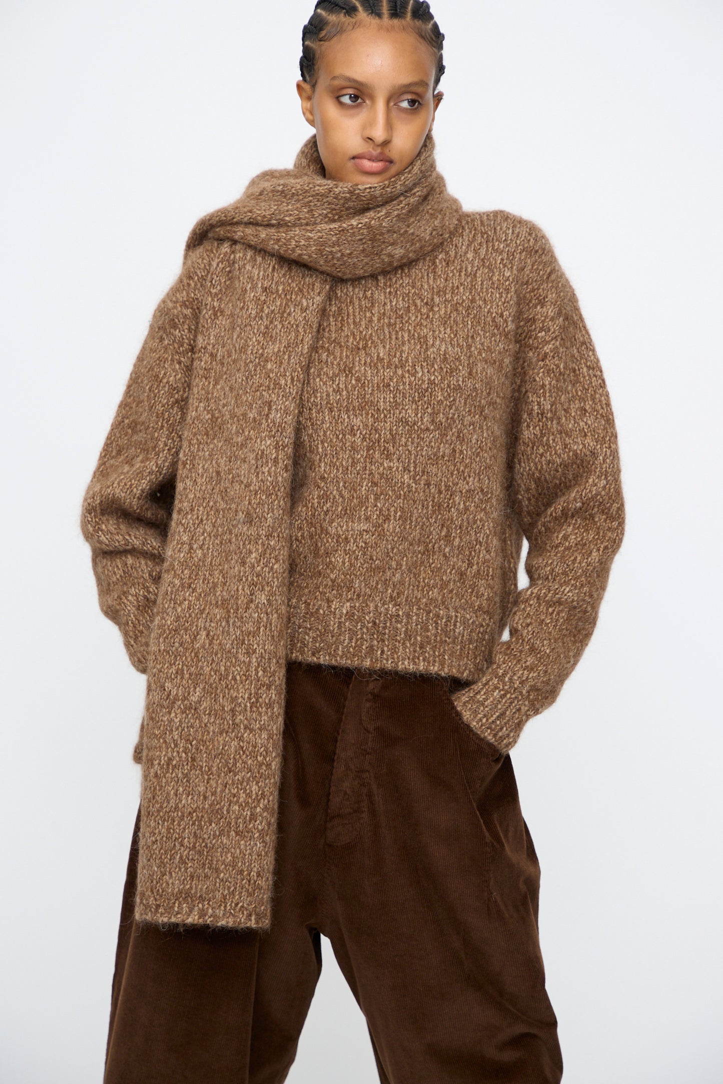 A person wears Cordera's Scarf in Brown Melange, styled with a brown knit sweater and matching corduroy pants, standing against a plain light background.