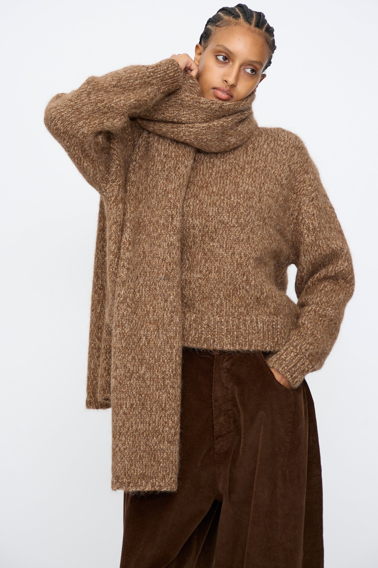 A person poses against a plain background, wearing brown pants, a chunky brown sweater, and the Cordera Scarf in Brown Melange with one hand on their head.