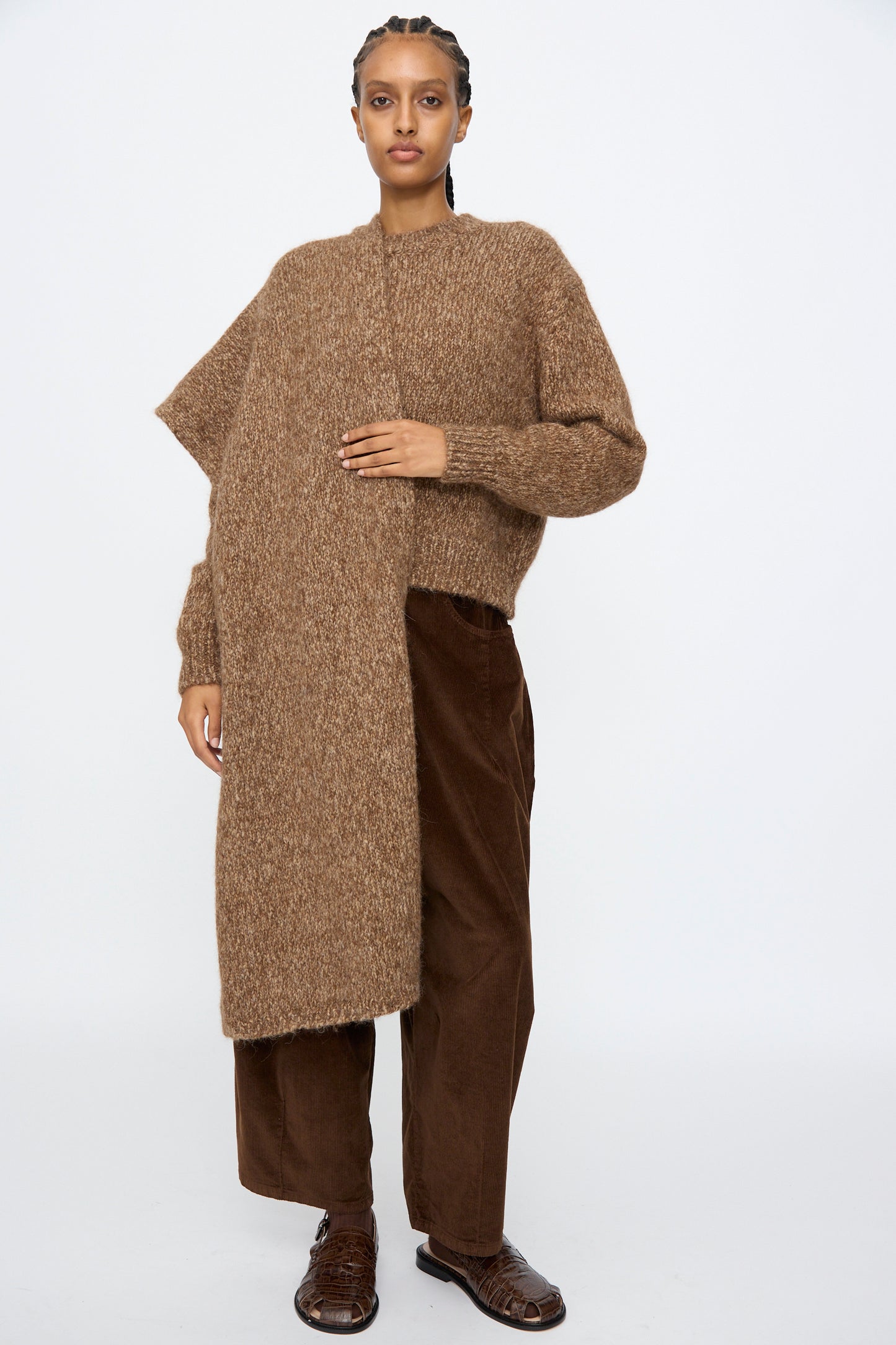 A person wears a brown asymmetrical knit sweater, brown corduroy pants, and brown shoes, styled with Cordera's Scarf in Brown Melange, standing against a plain white background.