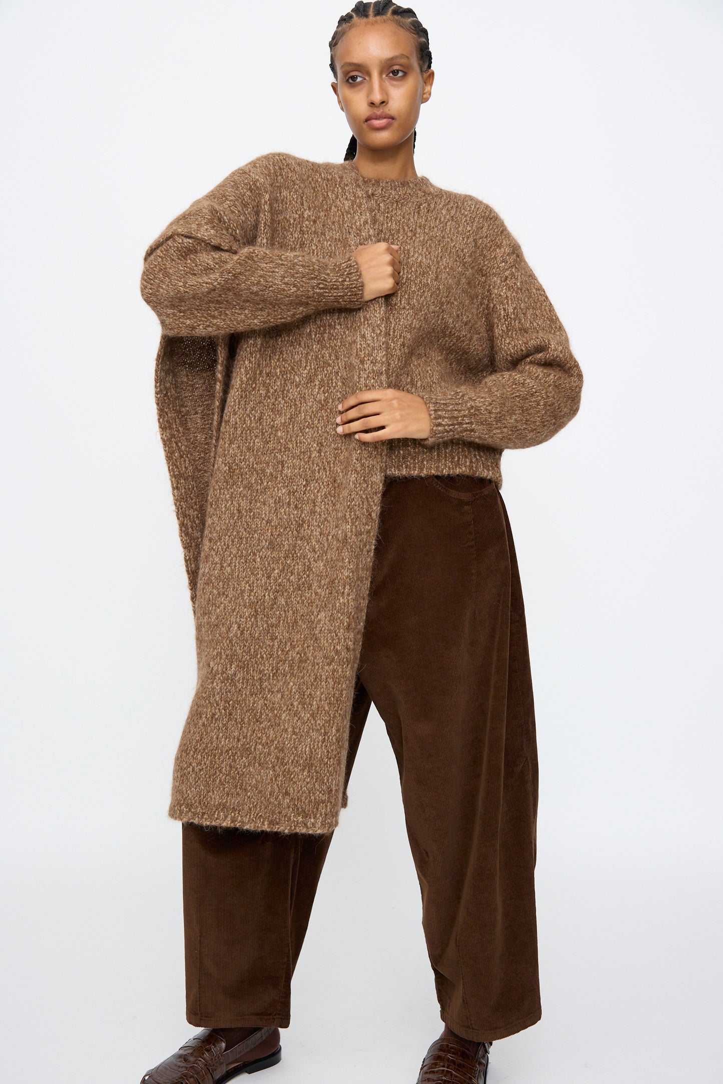 A person stands against a plain background wearing a loose brown knit sweater, matching brown melange wide-leg pants, brown shoes, and the Cordera Scarf in Brown Melange softly draped around the neck.