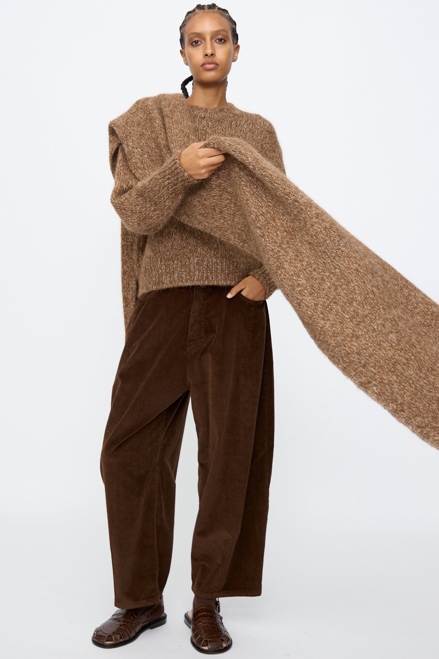 A person wears Cordera's Scarf in Brown Melange with a brown melange knit sweater, loose dark brown pants, and brown shoes, standing against a plain light background.