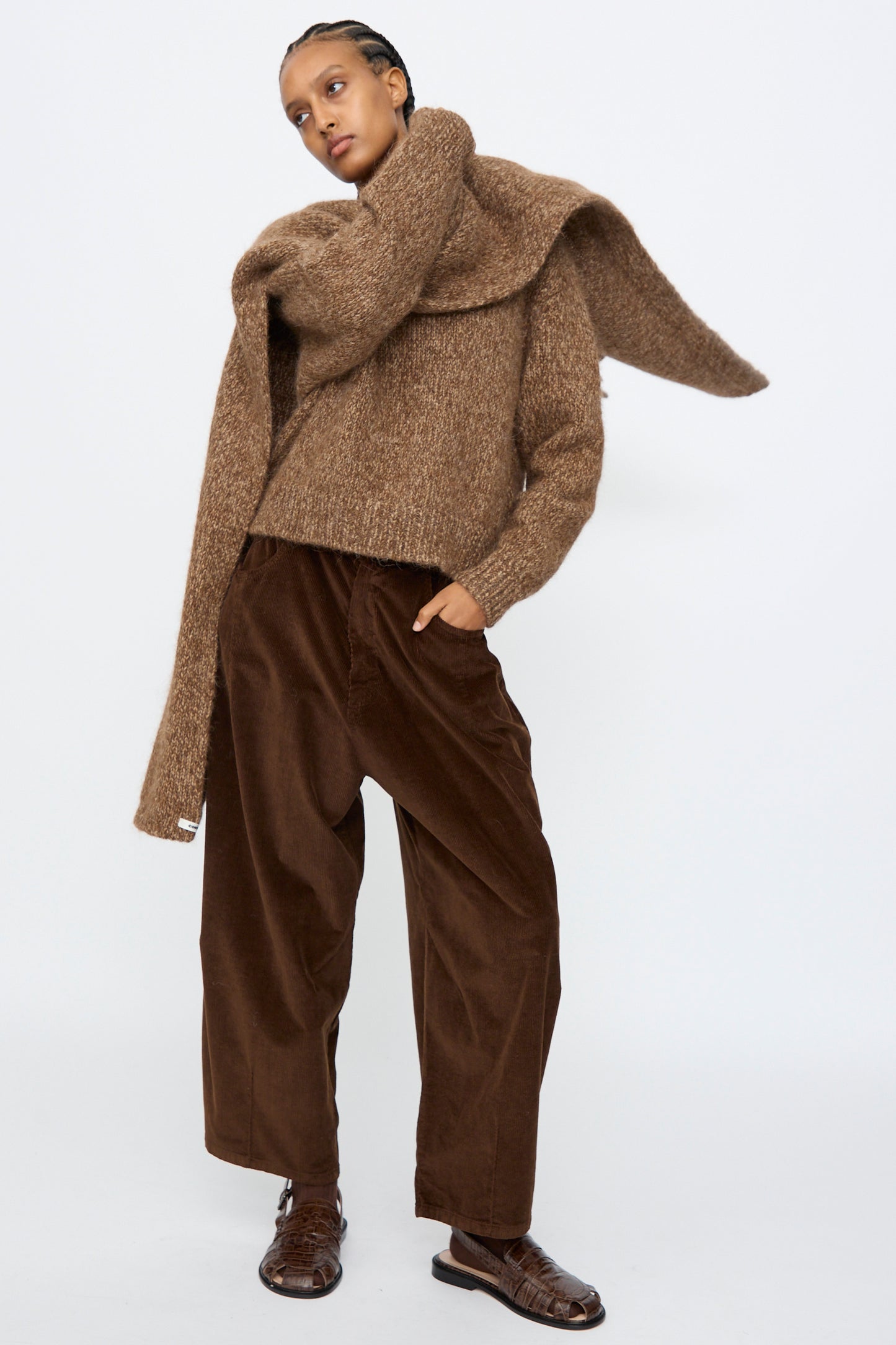 A person wears the Cordera Scarf in Brown Melange, styled with wide brown corduroy pants and brown shoes, standing against a plain white background.