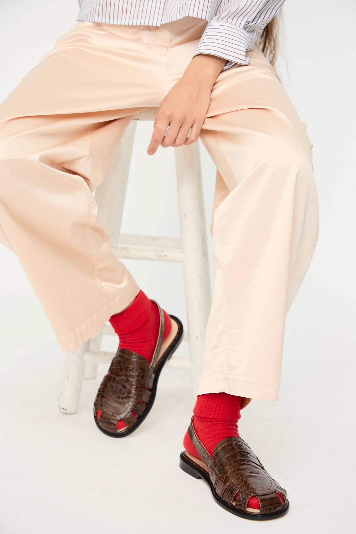 A person sits on a stool wearing beige pants, a striped shirt, bright red socks, and Cordera’s Slingback Loafer in Crocodile—brown loafers made in Spain—with one hand resting on their knee.