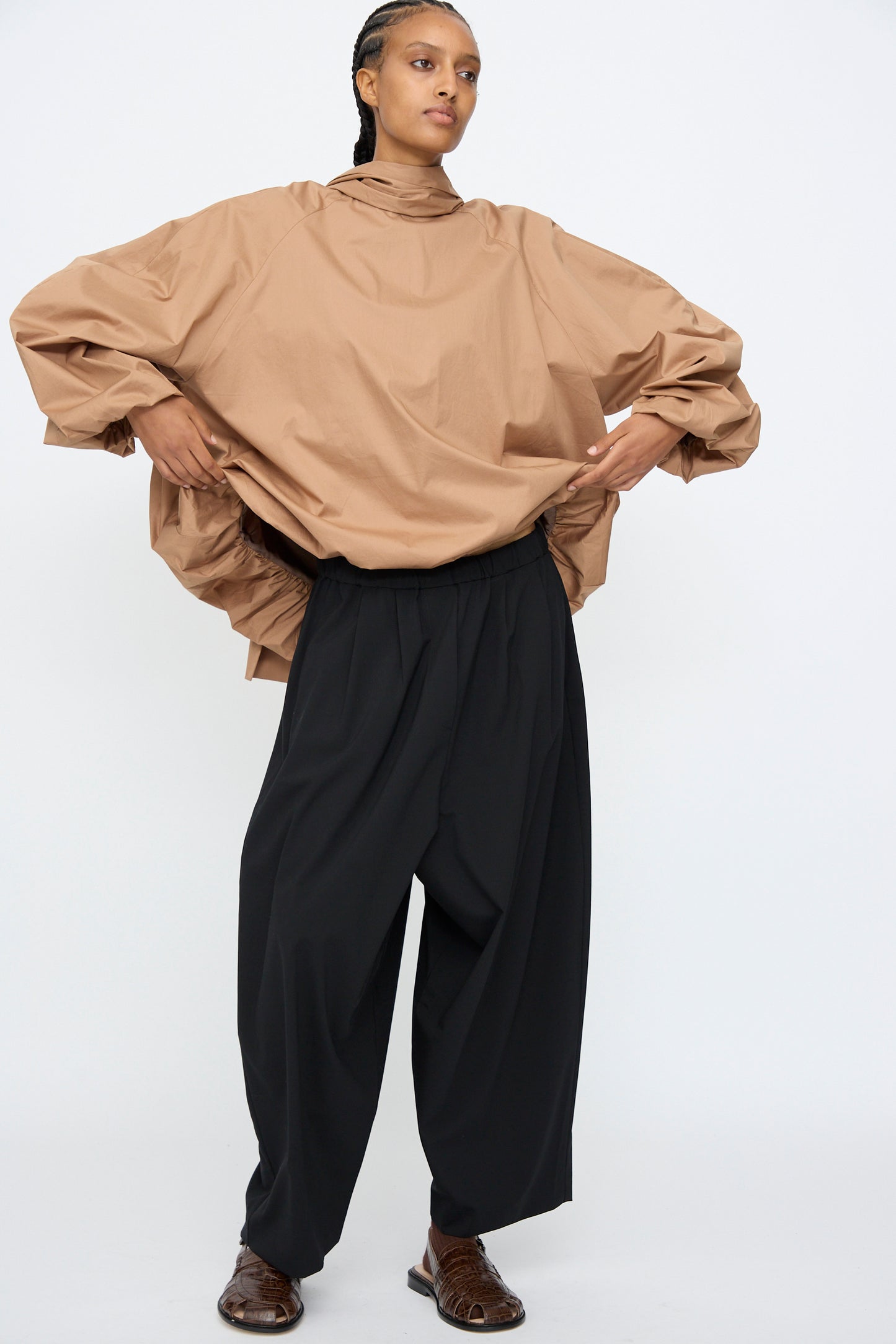 A person stands against a plain background wearing loose tan top, brown shoes, and Cordera’s Tailoring Maxi Pants in Black with an elastic waistband, arms bent outward.