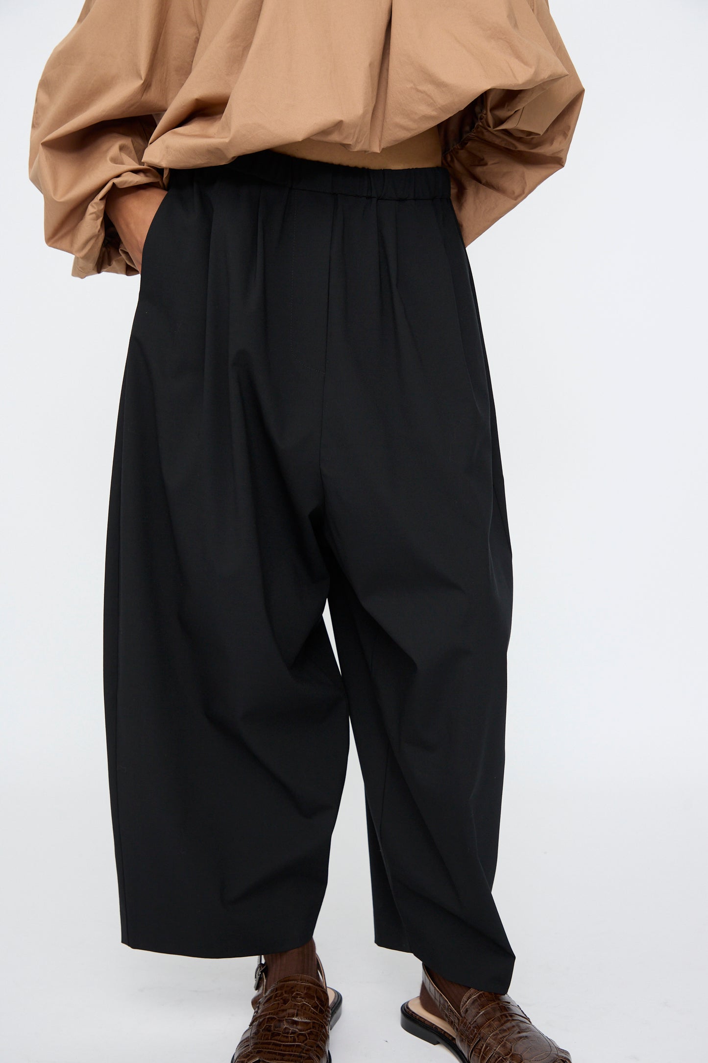 A person wearing Cordera Tailoring Maxi Pants in Black, paired with a loose tan shirt and brown textured shoes, stands against a plain light background.